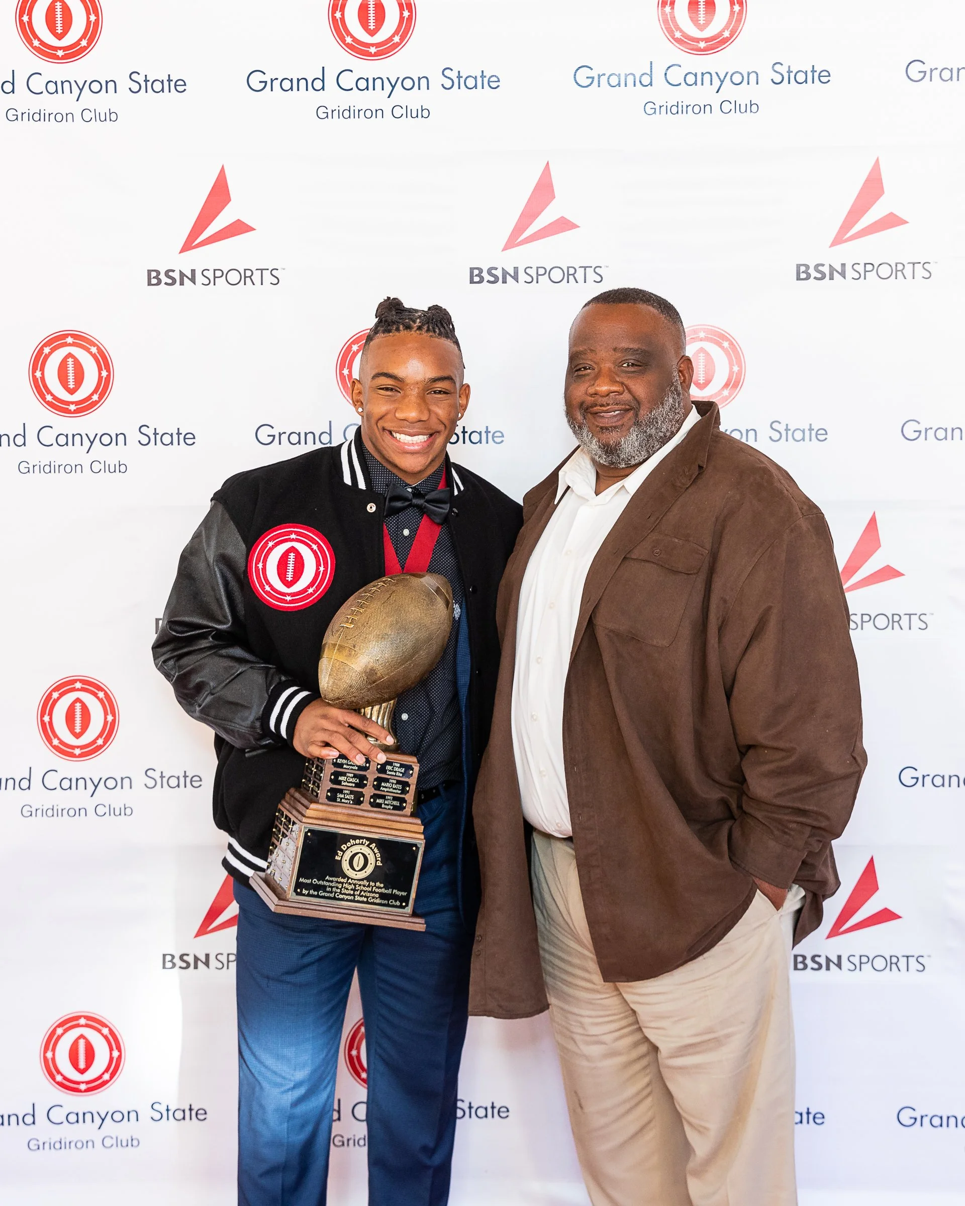 Two men standing together at an event, one holding a trophy shaped like a football. The background displays logos for Grand Canyon State Gridiron Club and BSN Sports.