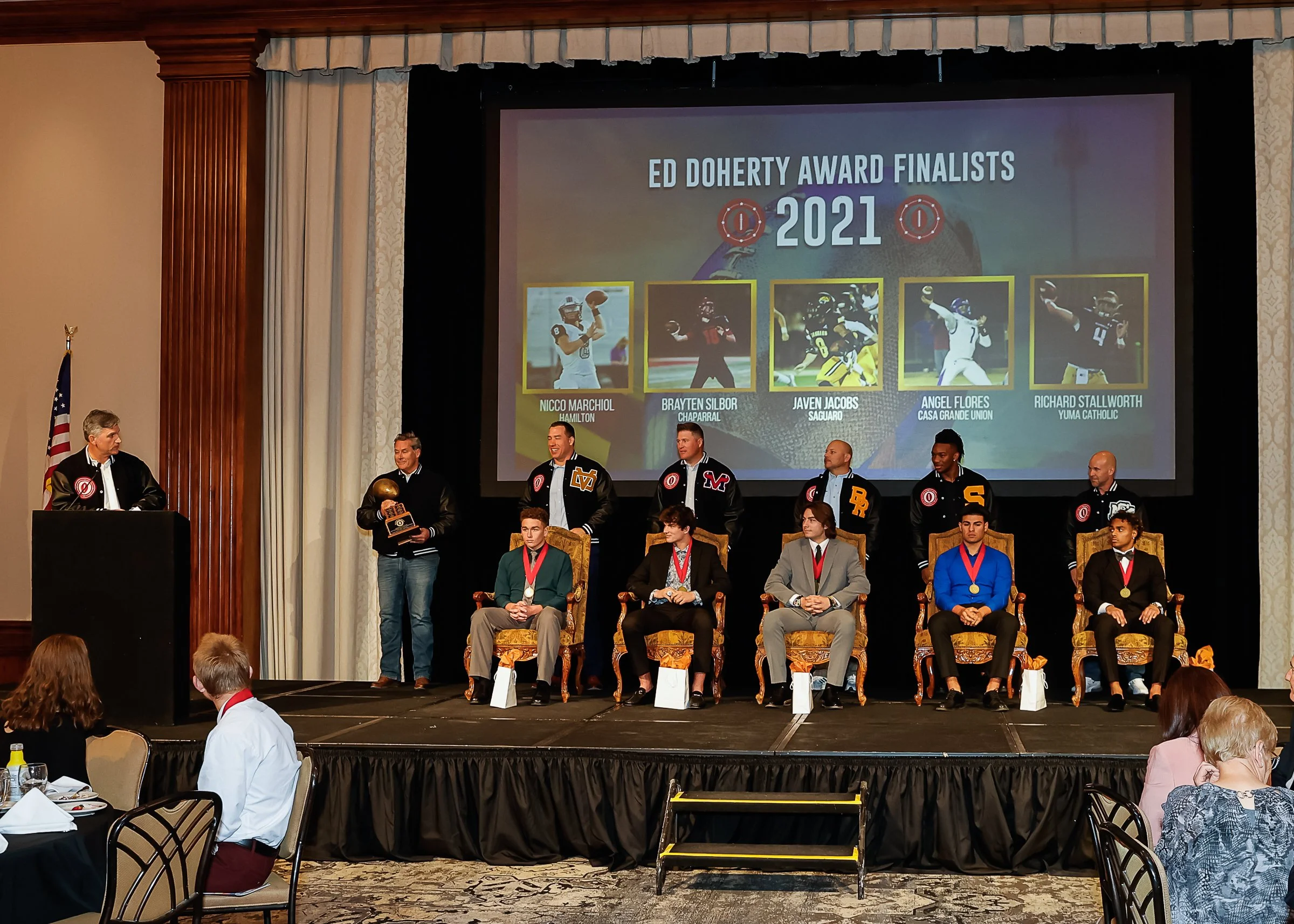 Award ceremony at a banquet hall with a stage where five athletes sit in ornate chairs, each wearing medals, and six men stand behind them. The large screen behind displays the text "Ed Doherty Award Finalists 2021" and photos of five football player