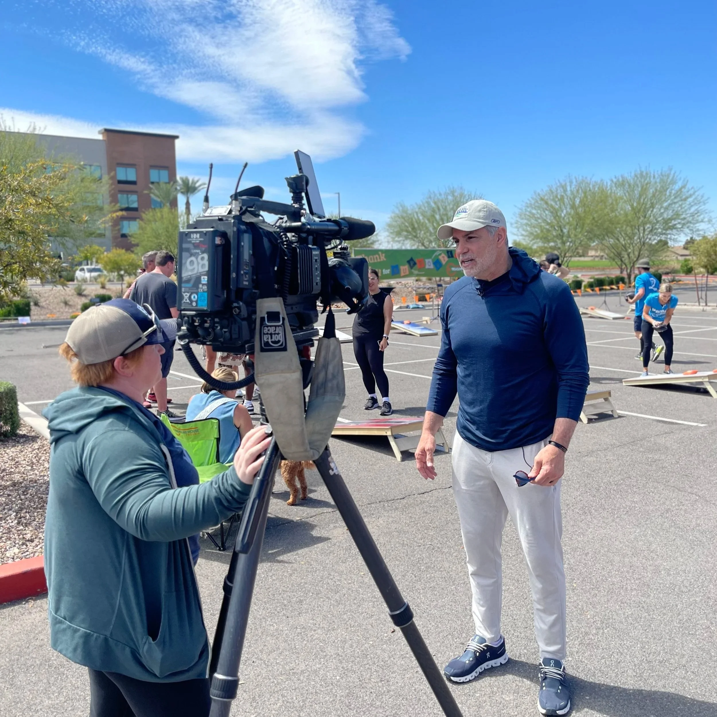 A man being interviewed by a woman with a camera in a parking lot, with people doing fitness activities on outdoor platforms in the background.