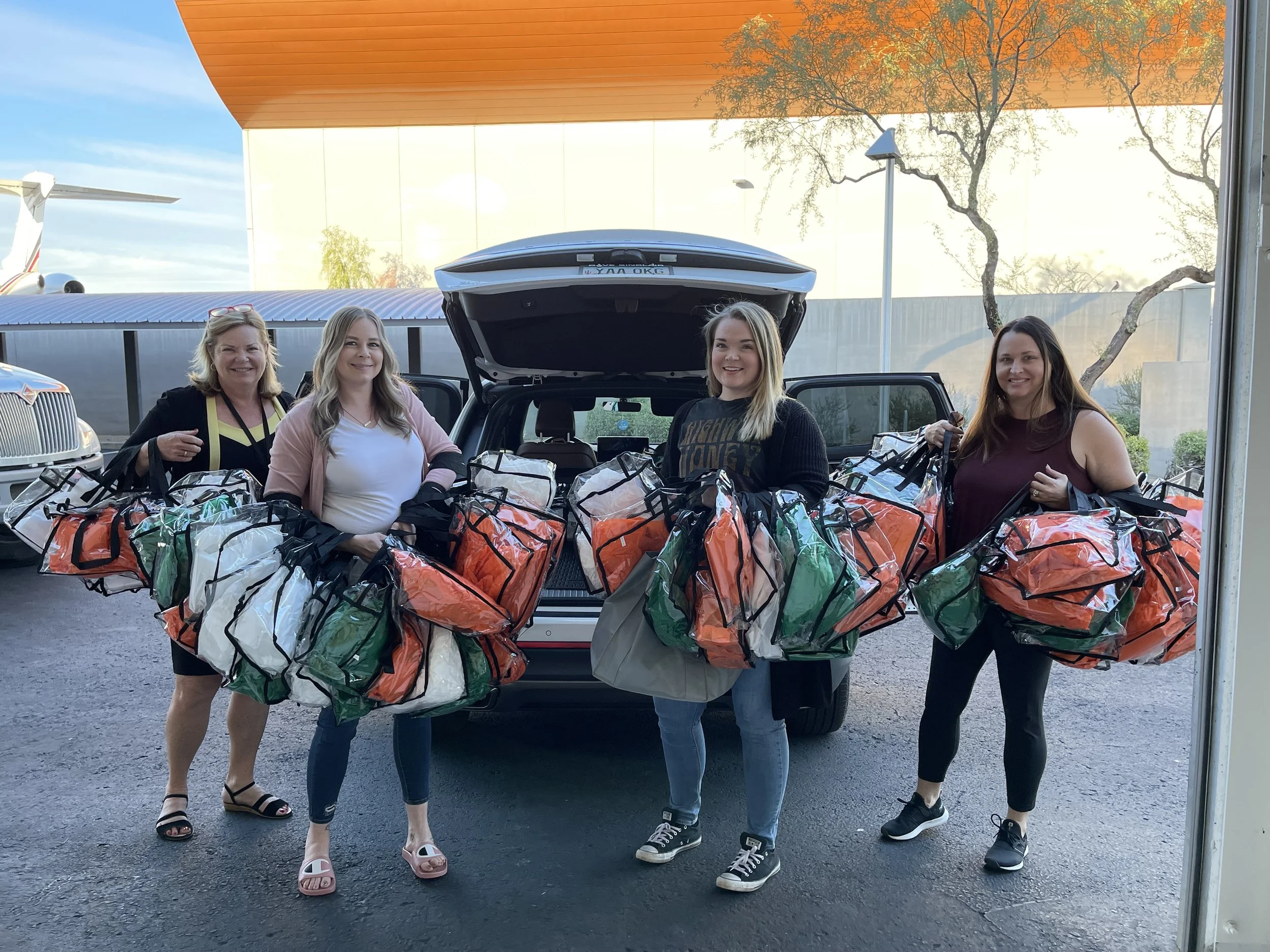 Four women standing outdoors behind a vehicle with its trunk open, each carrying multiple large paper bags filled with items, in a parking lot with a building and a tree in the background.