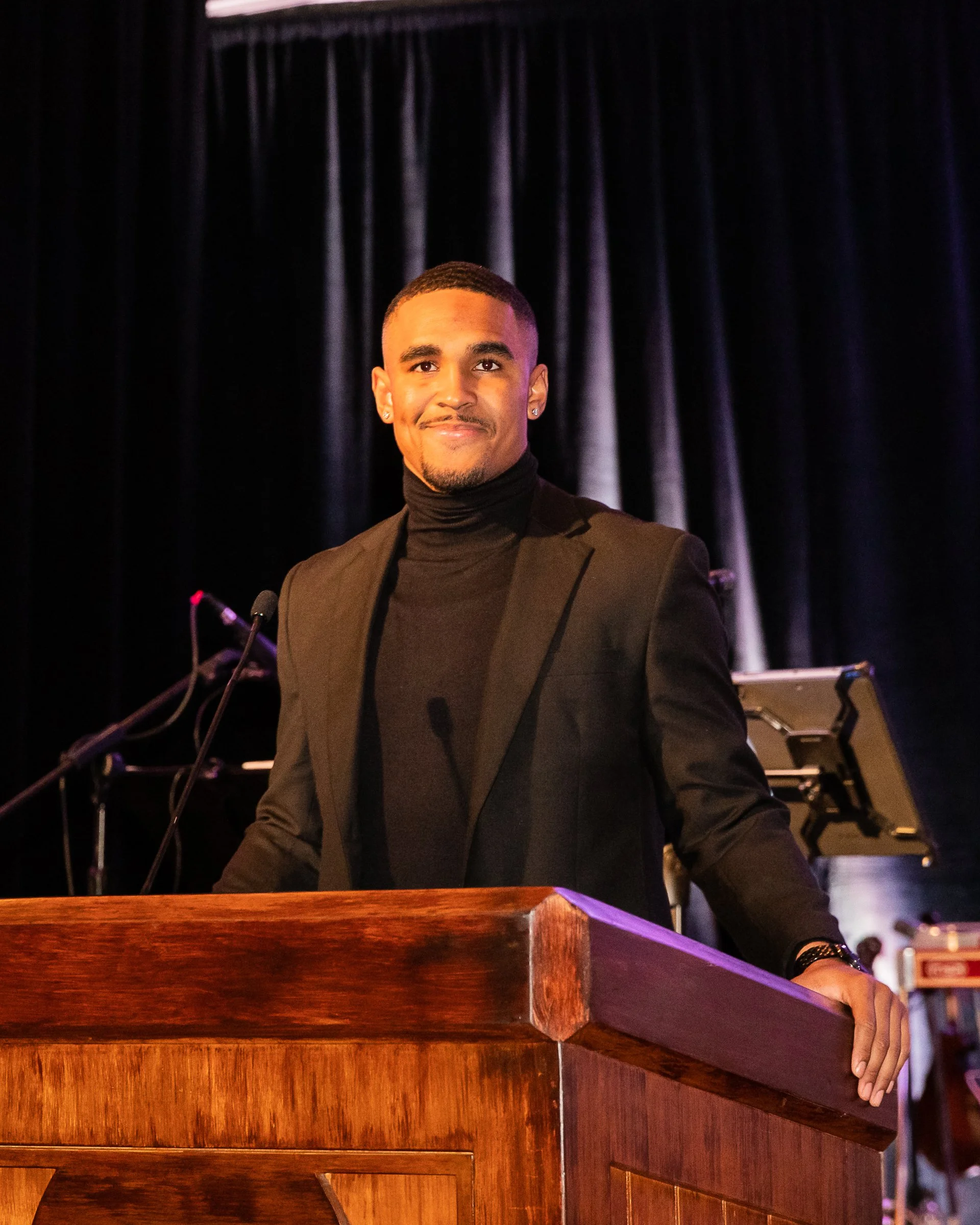 A man in a black turtleneck and blazer standing behind a wooden podium on a stage, with a dark curtain and some musical equipment in the background.