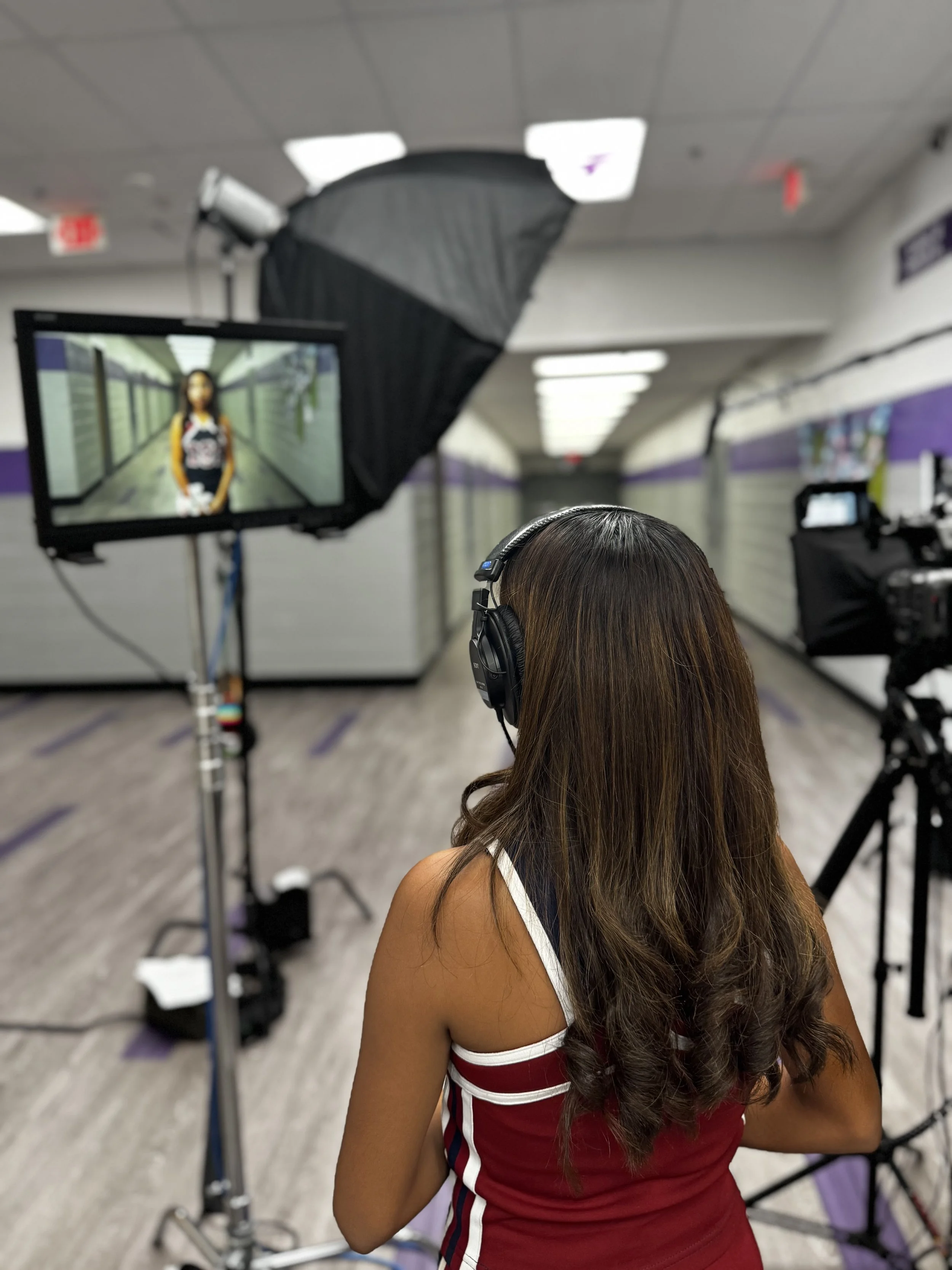 A woman with long brown hair wearing a red and white sports jersey is recording a video or photo in a hallway with purple walls and a gray floor. She is wearing headphones and facing a monitor that displays her image, with professional lighting and camera equipment set up around her.