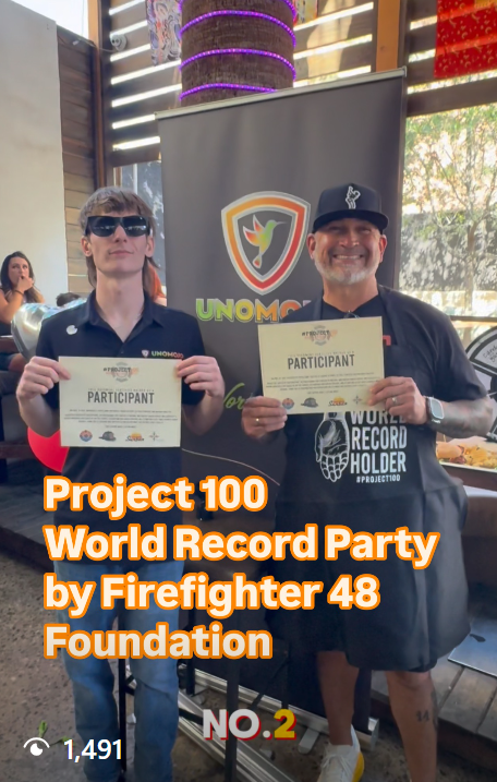 Two men stand together holding certificates with the event name 'Project 100 World Record Party' in front of a banner for UNOMIC. One man is wearing sunglasses and a dark shirt, the other smiling man is wearing a black cap, gray shorts, and a black T-shirt that says 'World Record Holder.'