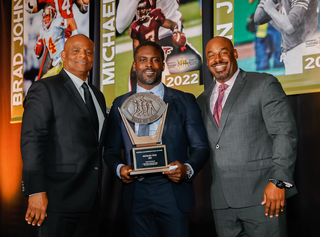 Three men in suits standing on a stage, one holding an award plaque. The background features sports banners and images of football players.