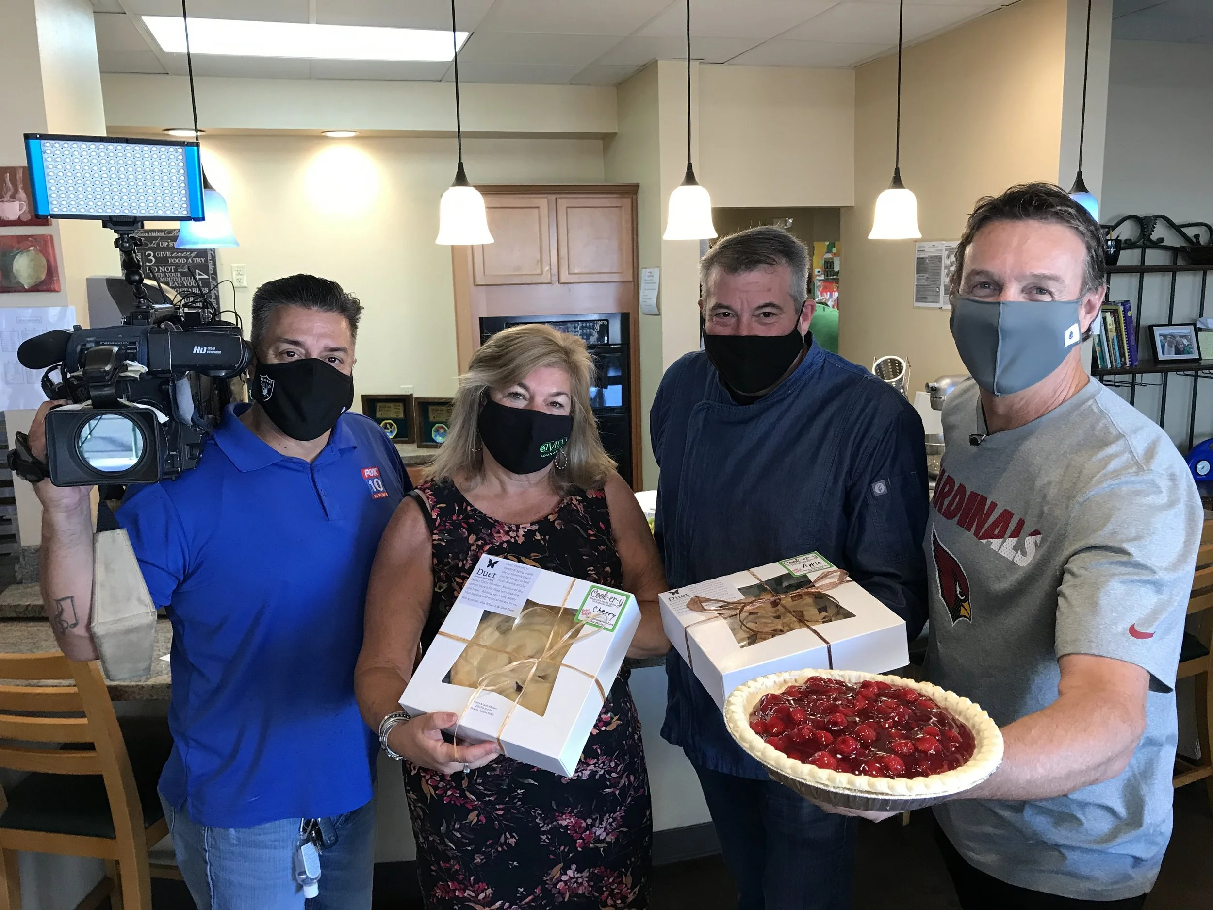 Four people, wearing masks, hold boxes and a pie, standing in a restaurant or cafe. A camera operator records the scene.