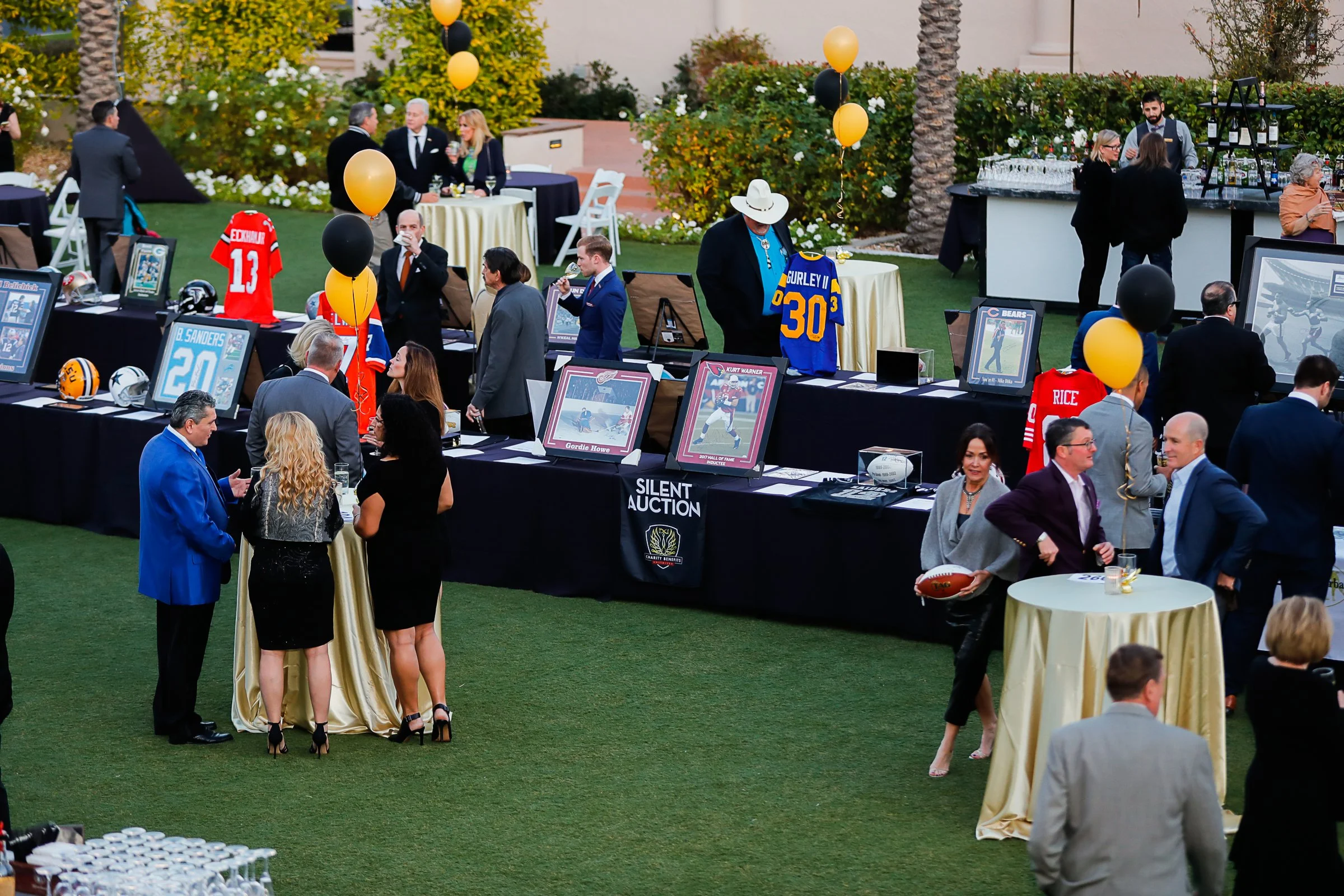 A silent charity auction event taking place outdoors on a grassy area, with tables displaying sports memorabilia, framed photos, and NFL jerseys. Attendees are mingling, dressed in business or semi-formal attire, with some holding drinks. Balloons de