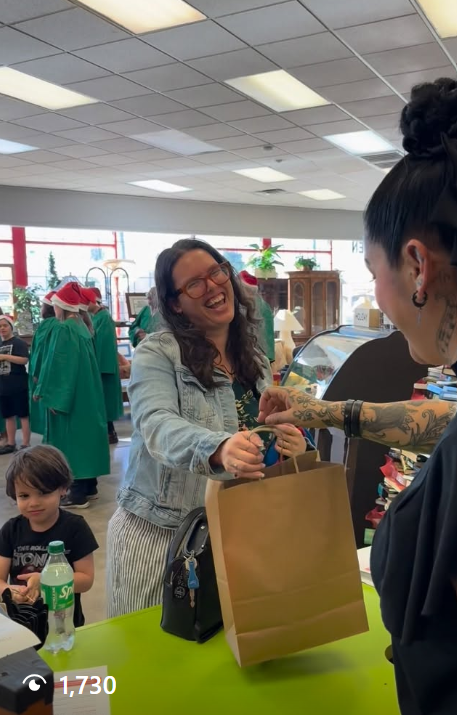 A woman with glasses and long hair is smiling and receiving a brown paper bag from a cashier at a store checkout counter, with other people and Christmas decorations in the background.