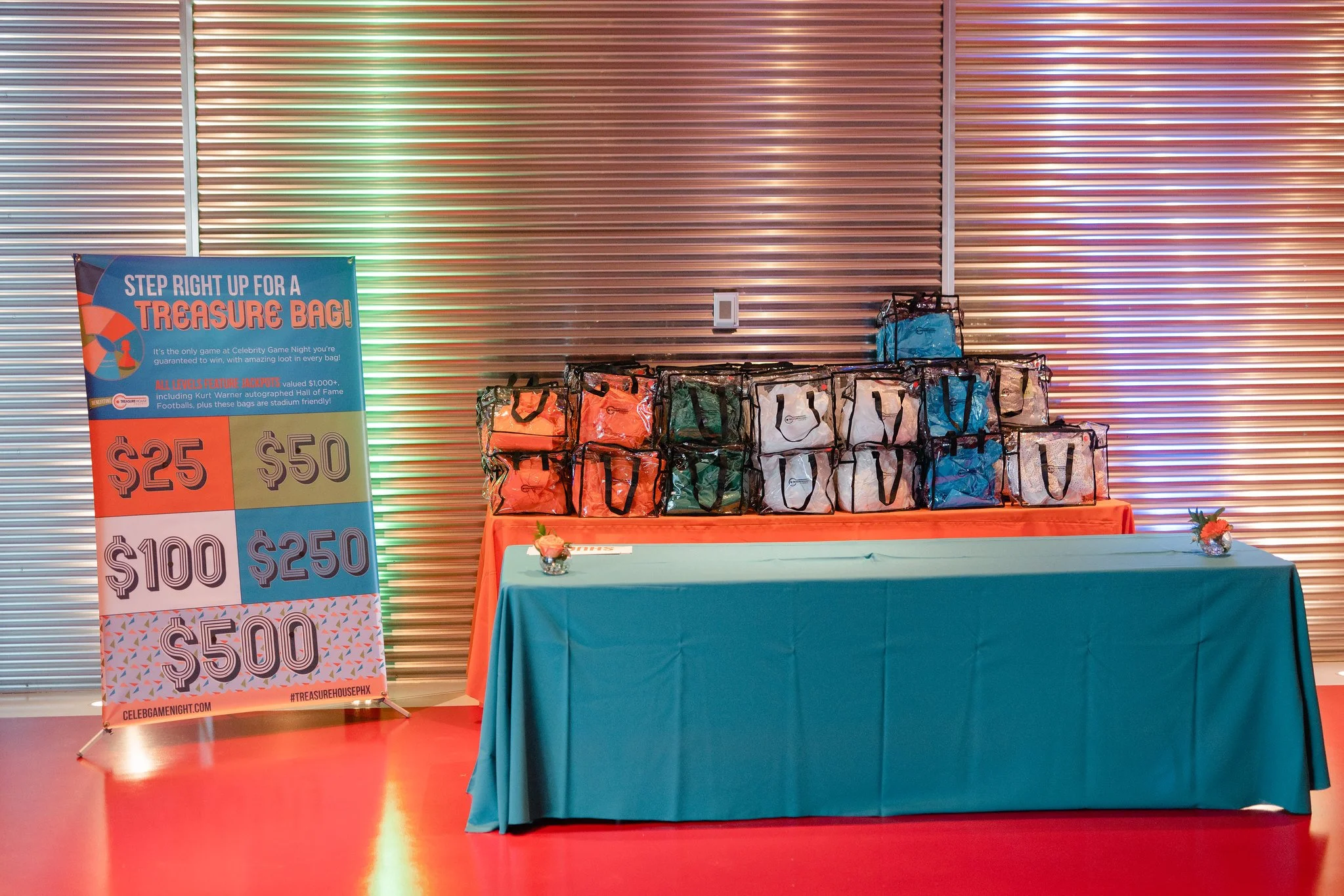 Table with multiple prize bags for a game at an event, with a sign listing cash prize amounts from $25 to $500, and a vase with flowers on each end of the table.