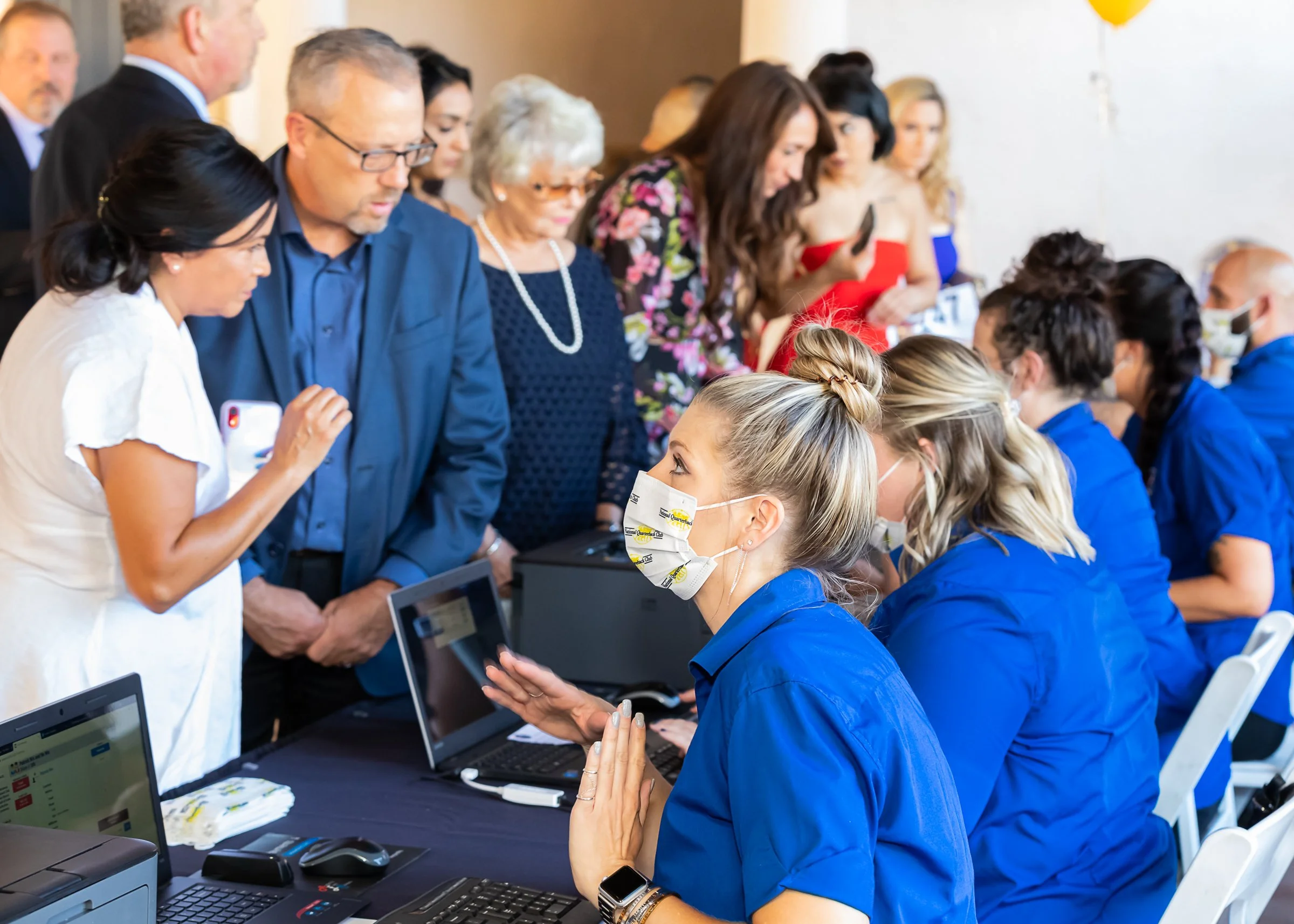 People gathering at a registration or check-in desk, with some individuals wearing blue uniforms and masks, while others stand in line or look on, at a busy indoor event.