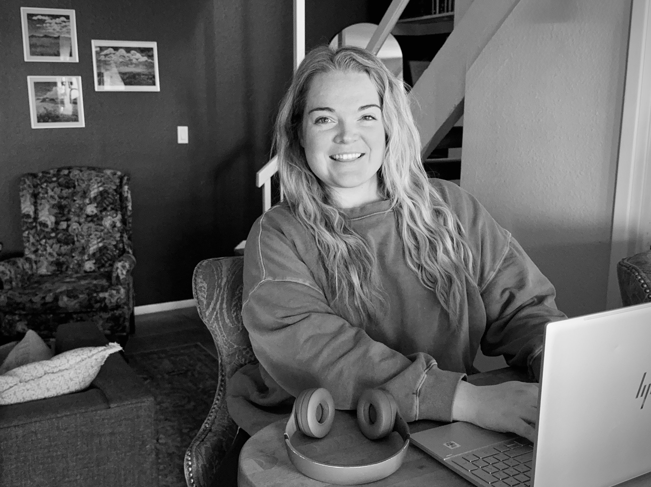 A smiling woman with wavy blonde hair sitting at a table with a laptop and headphones, in a cozy living room with framed pictures and patterned furniture.