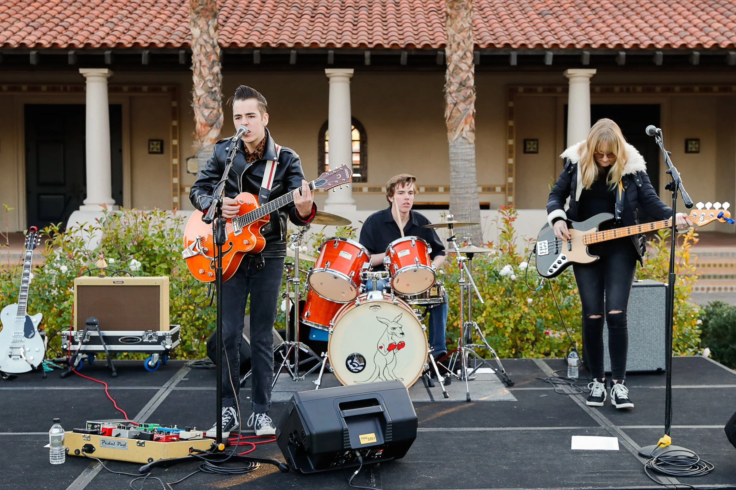 Three musicians performing on an outdoor stage. The lead singer is playing an orange guitar and singing into a microphone. To his right, a woman is playing a black bass guitar. In the background, a drummer is playing a drum set decorated with a drawi