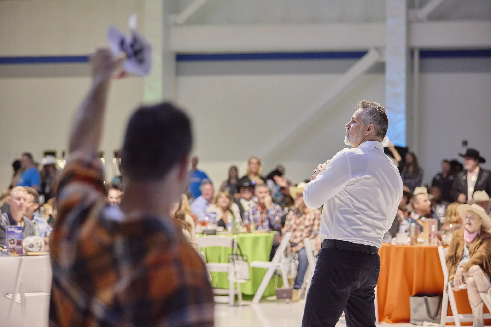A man with gray hair, dressed in a white shirt and black pants, speaking to a crowd at a large indoor event with round tables and yellow, green, and orange tablecloths.