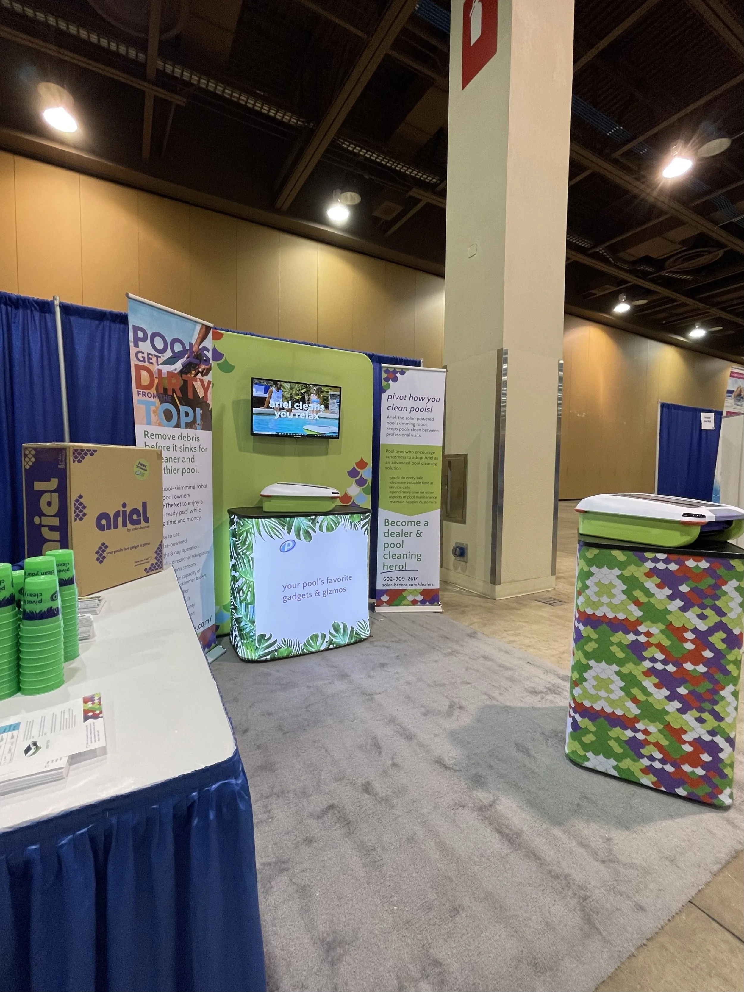 Trade show booth promoting pool cleaning gadgets and Gizmos with banners and displays, including a television screen, table with green cups, and colorful equipment covers.