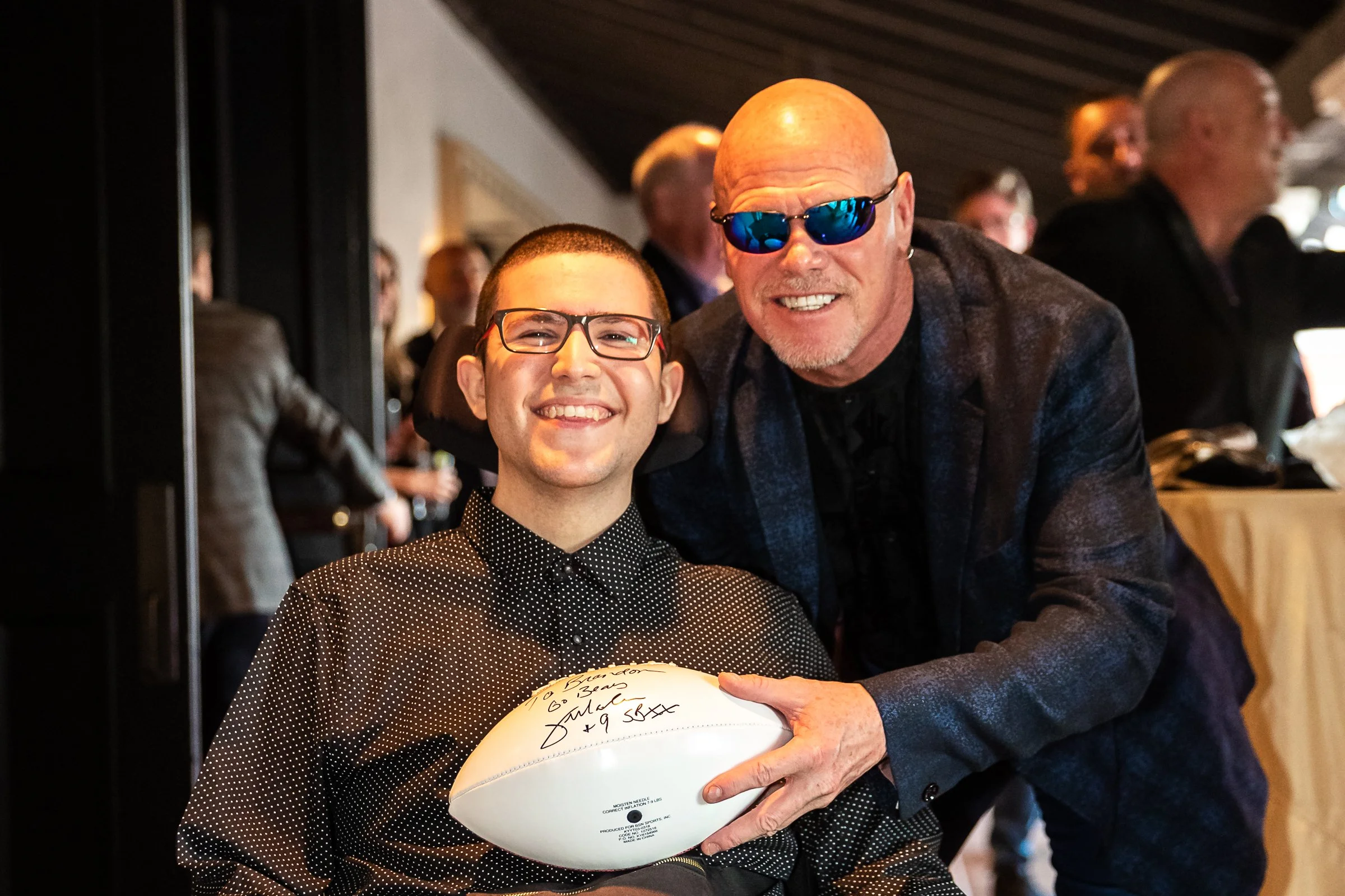 A young man with glasses, smiling, holding a signed football, and an older man with sunglasses, smiling, leaning in close at a social event.
