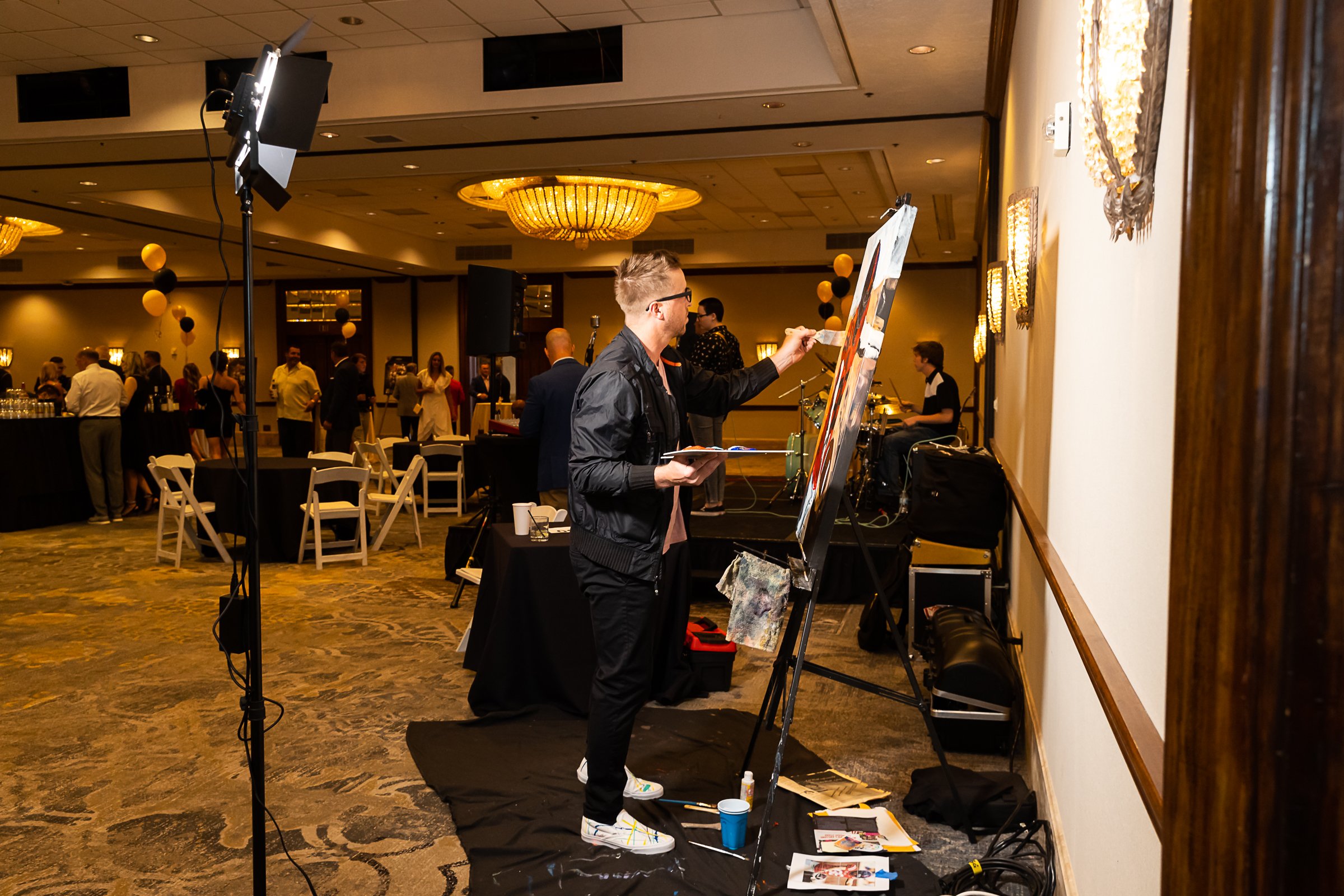 An artist painting on a canvas at an indoor event with people socializing and a band playing in the background.