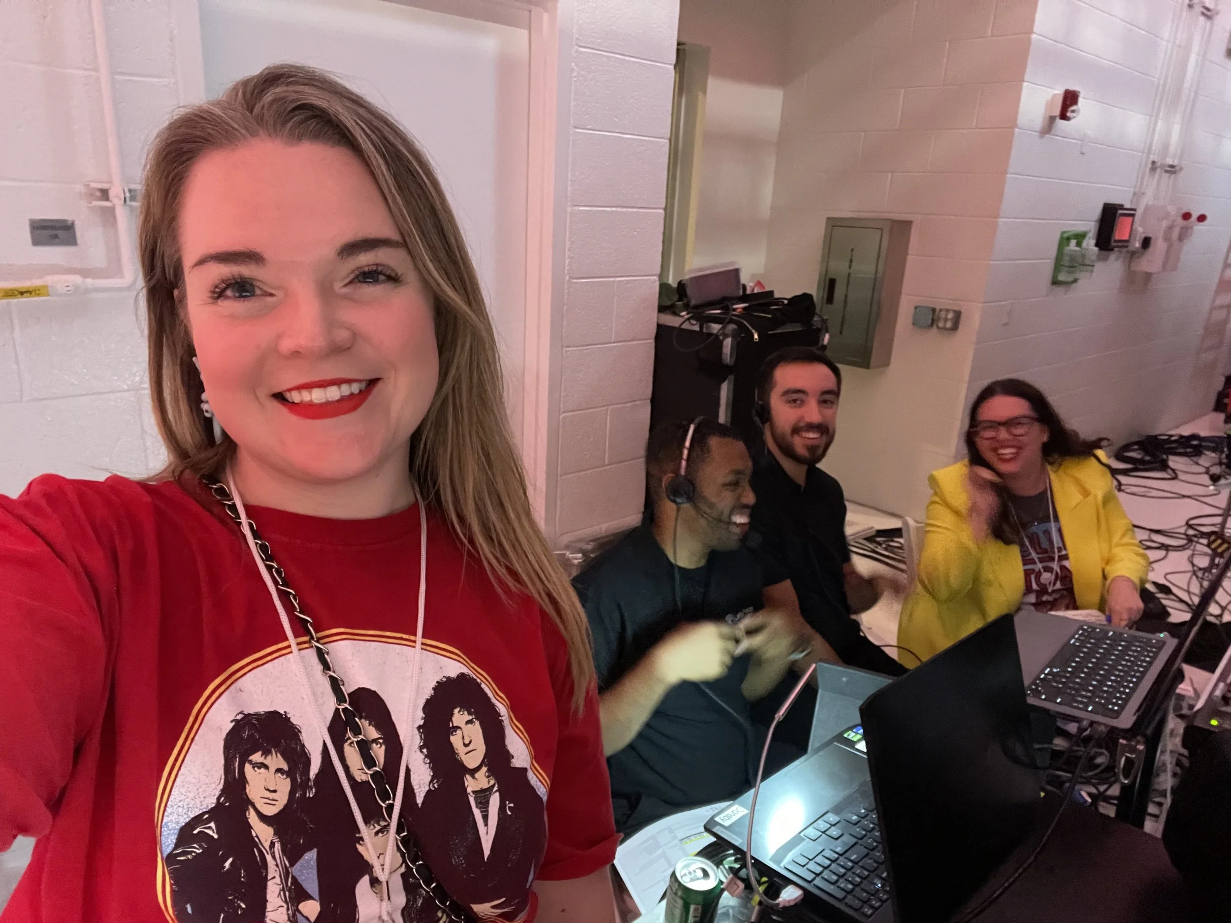 A woman with long blonde hair wearing a red T-shirt with a graphic of four people, smiling at the camera in a room with four other people sitting at a table with laptops and equipment, laughing and talking.