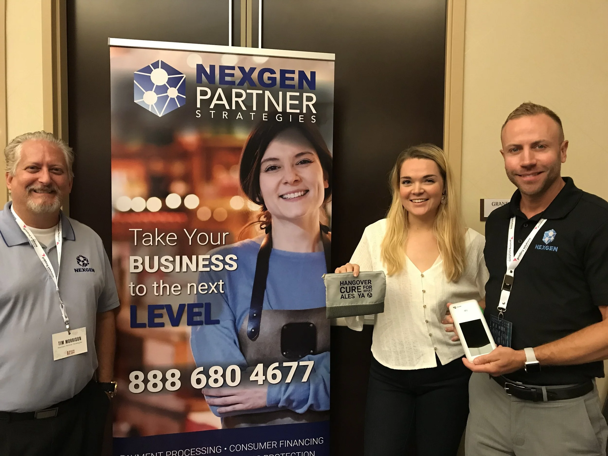 Three people posing next to a NEXGEN Partner Strategies banner at a business event. The banner features a smiling woman and the text 'Take Your Business to the Next Level' along with a phone number.
