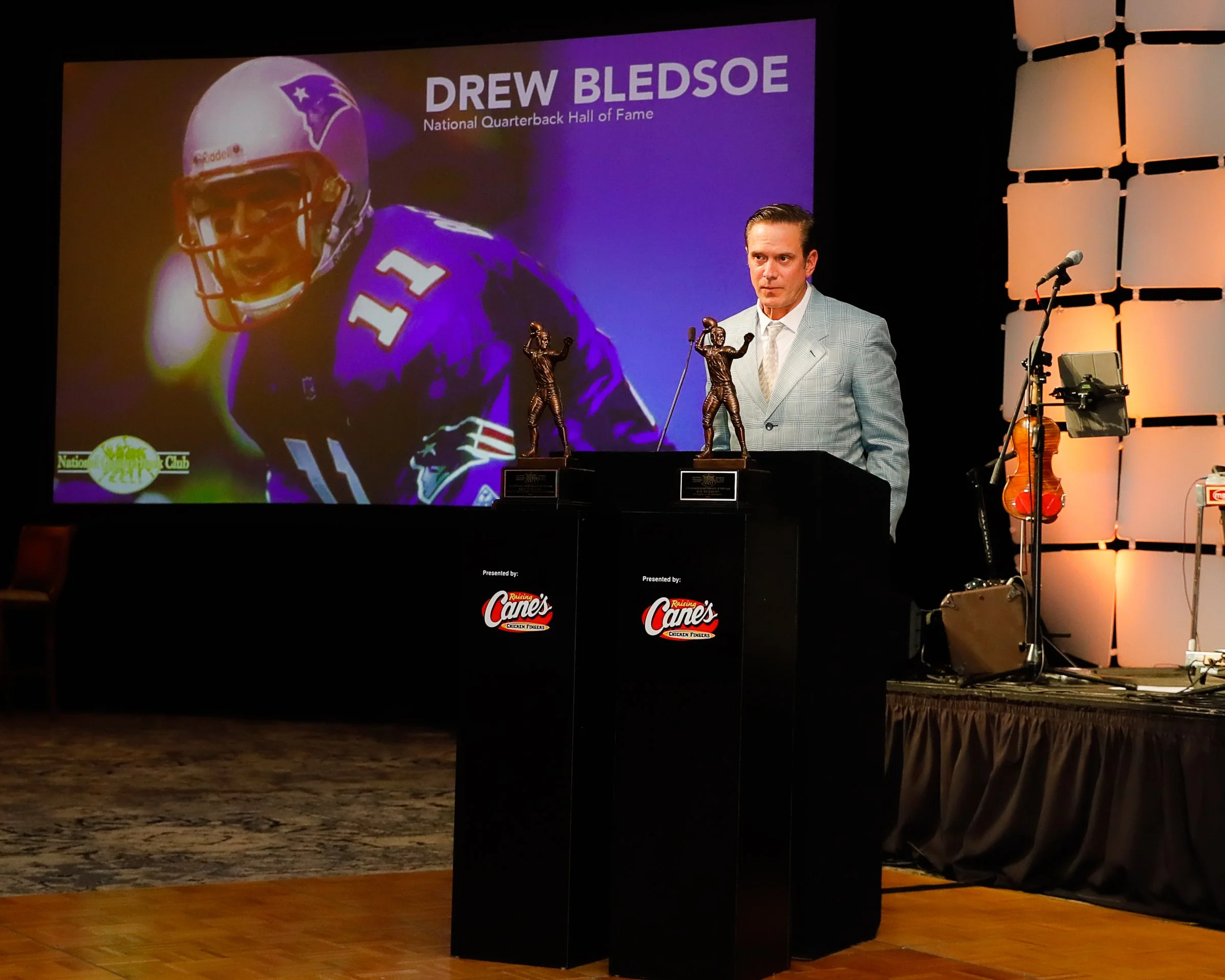 Drew Bledsoe at a Hall of Fame induction ceremony, standing behind a podium with football trophies, in front of a large screen displaying his image and name.