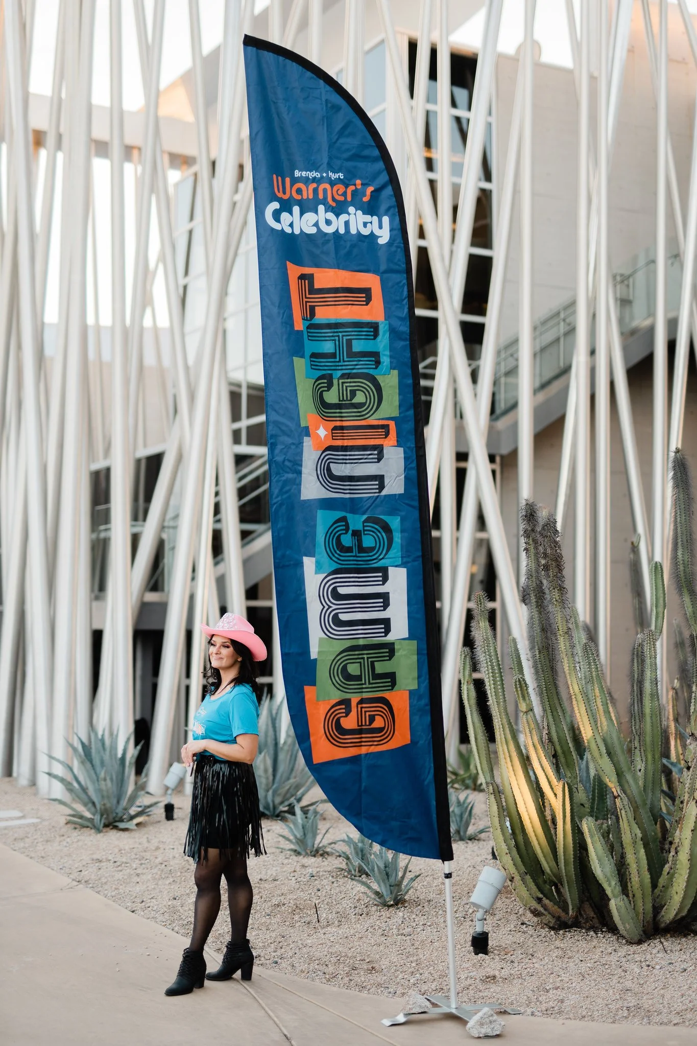 A woman wearing a pink cowboy hat, a blue t-shirt, a black fringed skirt, and black high-heeled boots standing next to a large blue banner that reads 'GALLERY' in colorful letters. The banner has additional text indicating an event called 'Brenda + K