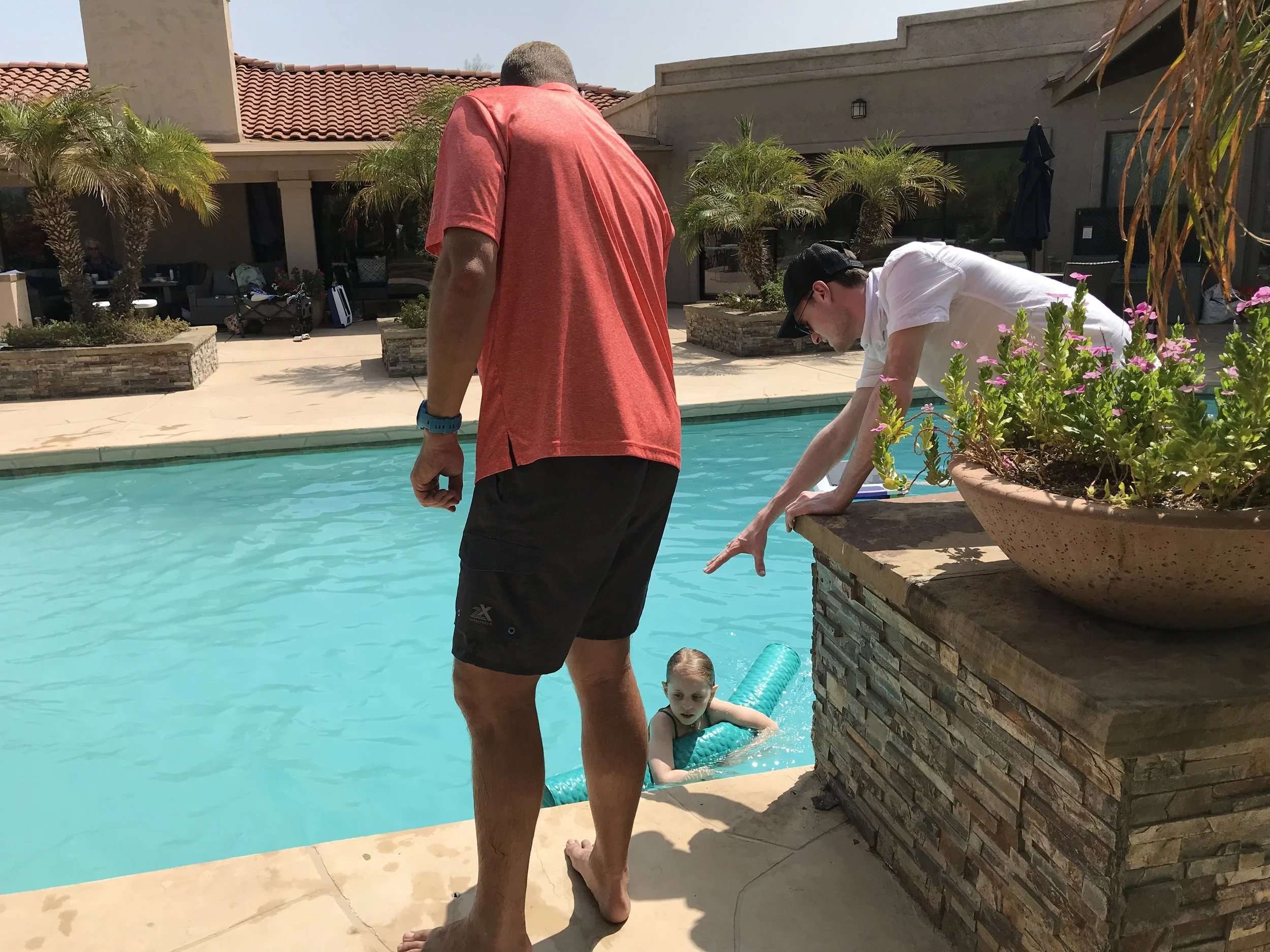 Two men stand at the edge of a swimming pool, observing a young girl in the water. The girl has a pool noodle, and one man is leaning over the pool, pointing at her. The setting is a sunny backyard with potted plants and patio furniture.