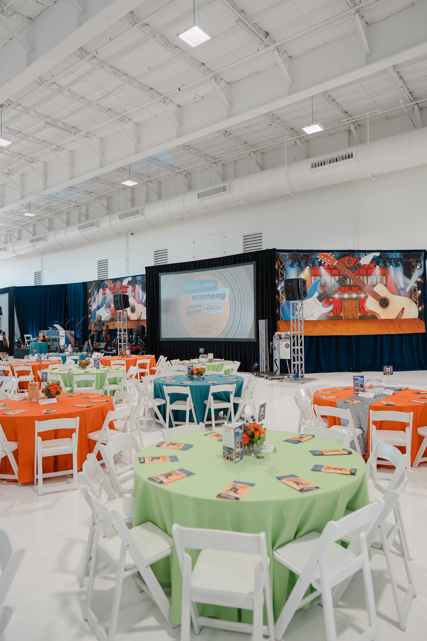 Event space with round tables covered in colorful tablecloths and white chairs, a stage with a large screen, musical instruments, and bright lighting in a spacious hall.
