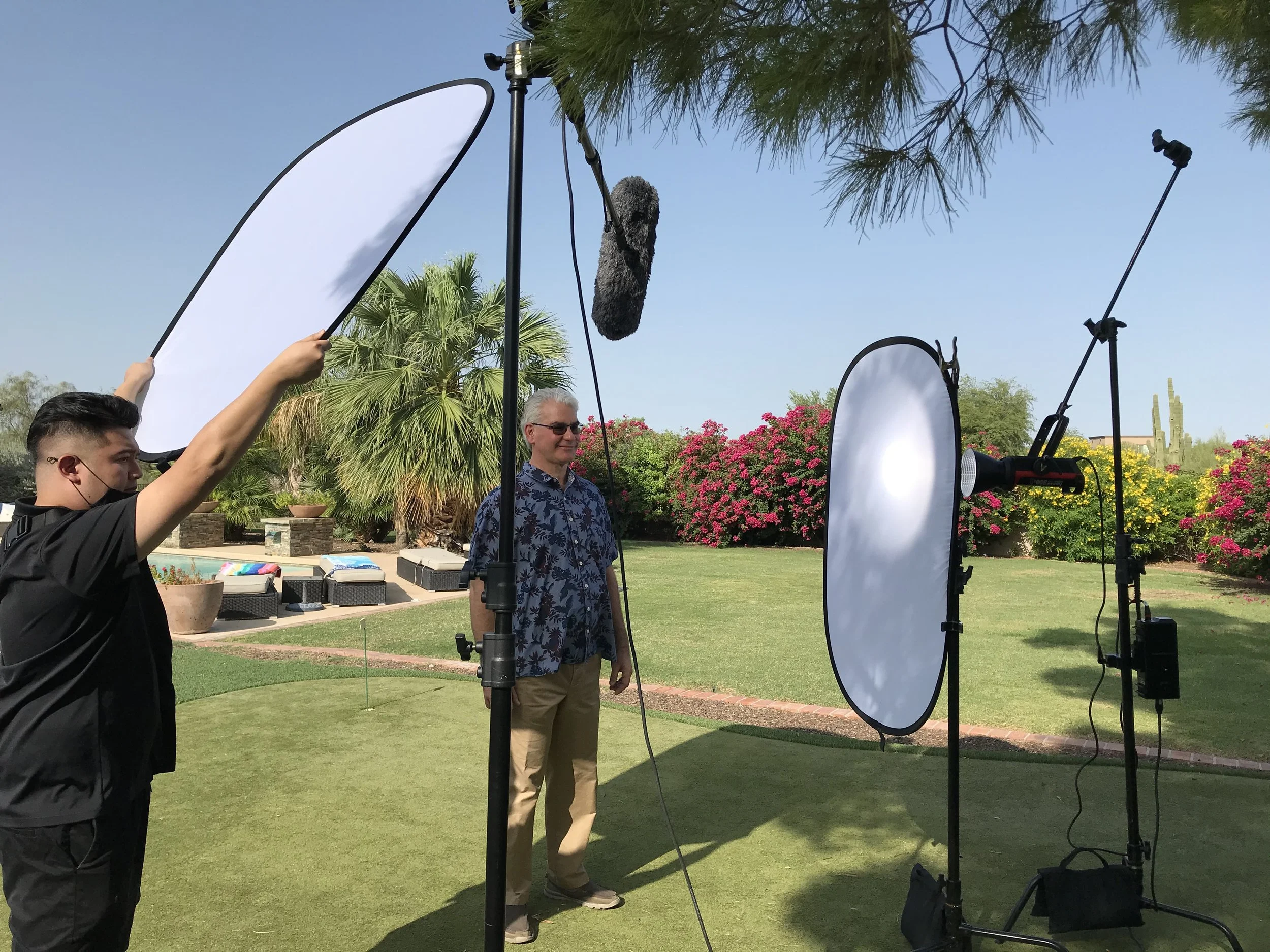 Filming a man outdoors with lighting equipment, large reflector, and a sound boom, in a garden with greenery, palm trees, and flowering bushes.