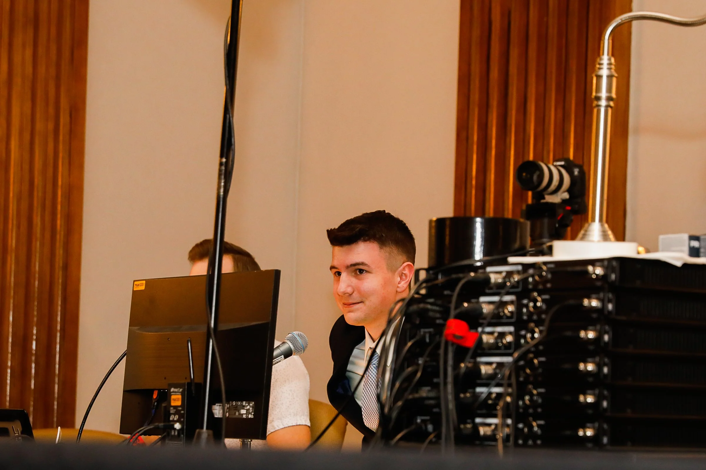 A young man sitting at a conference table, smiling, with a microphone, computer monitor, and audio/video equipment in front of him, in a room with wooden paneling.