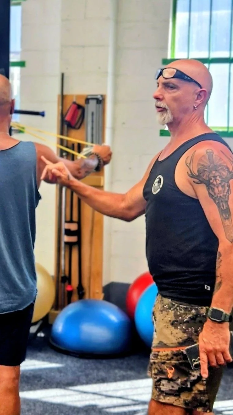 A muscular man with glasses on his forehead, tattoos, and a beard, standing in a gym and instructing or talking to another person during a workout session. The gym has exercise equipment and colorful exercise balls in the background.