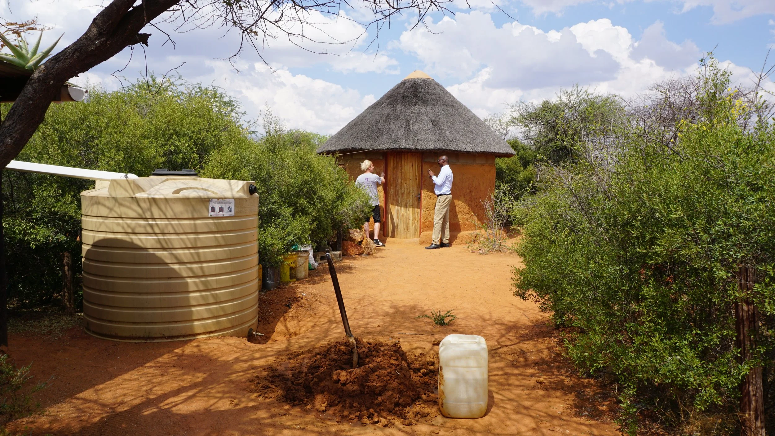 Two people standing outside a traditional round mud and thatch hut, surrounded by greenery and blue sky with clouds. One person is using a phone, and the other is near the door of the hut. In the foreground, there is a water tank, a small pile of dir