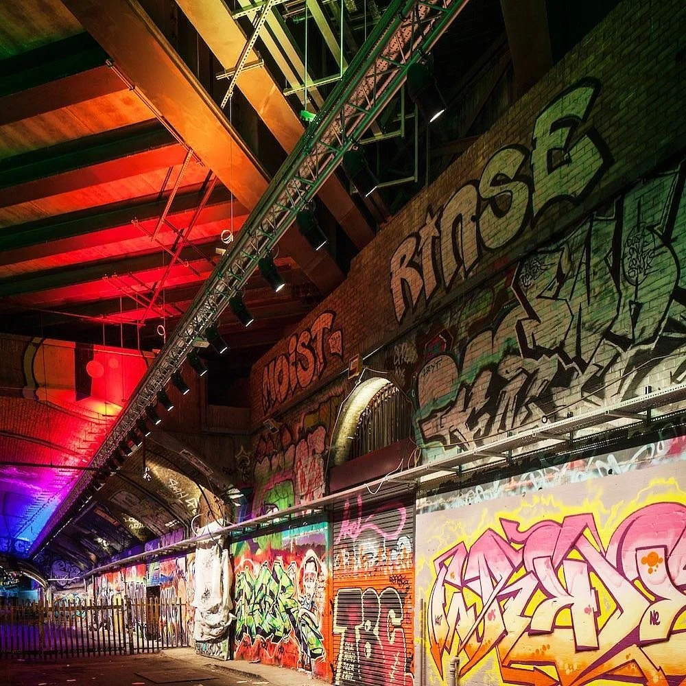 Graffiti art on brick walls in an urban tunnel under a bridge, illuminated with colorful lighting.