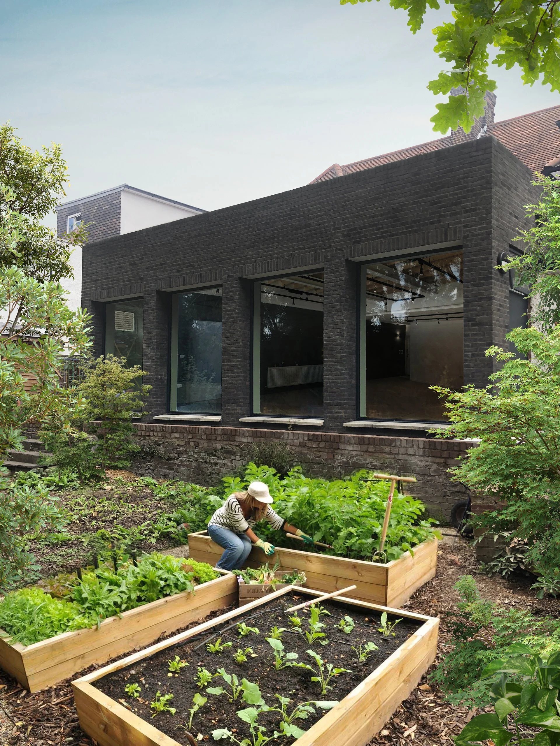 A woman gardening in a backyard farm with raised wooden beds filled with leafy greens and vegetables, a modern black brick building with large windows in the background, and surrounding trees and plants.