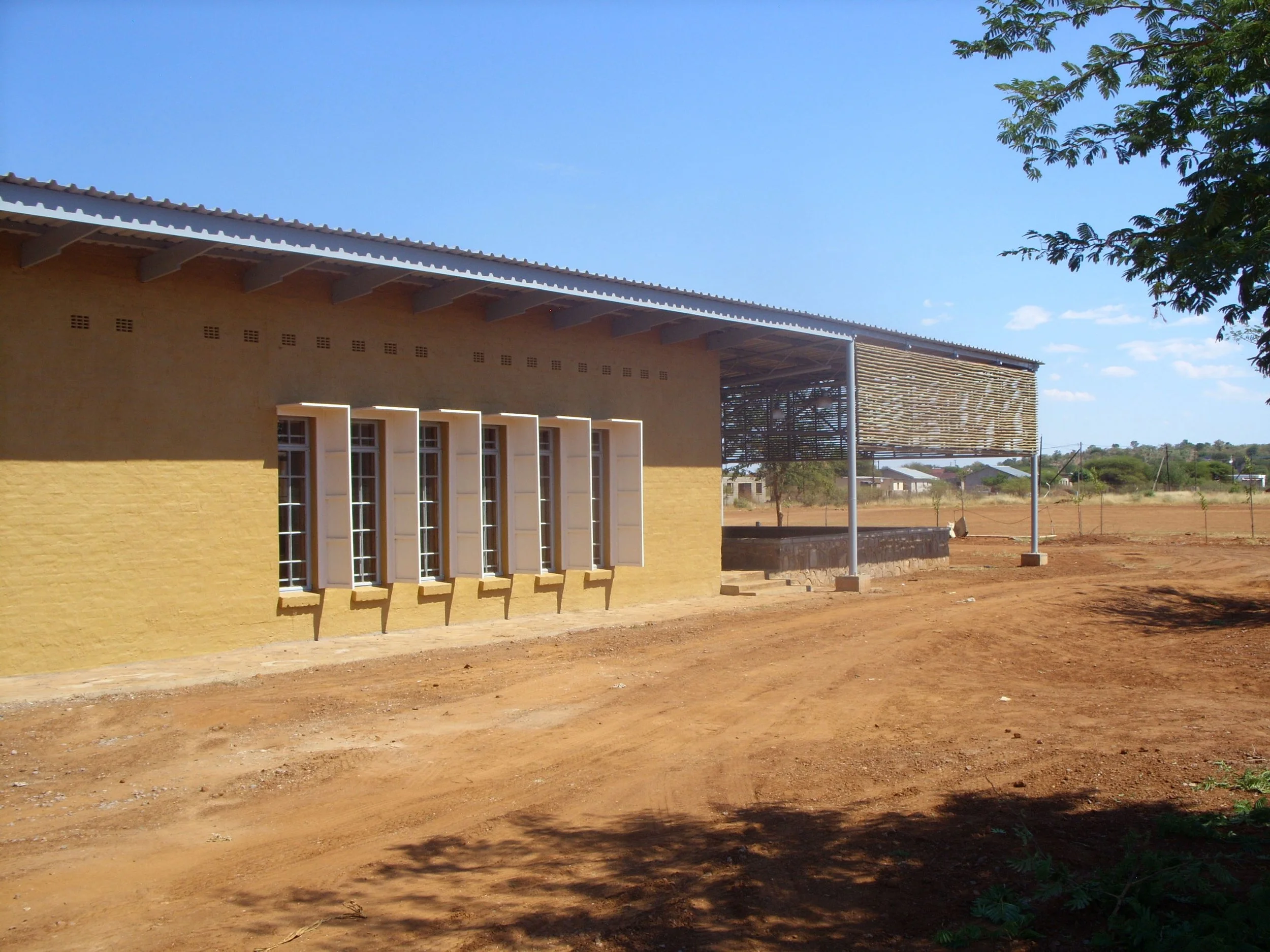 A yellow building with open white window shutters, a corrugated metal roof, and an outdoor shaded area with a wooden slat wall, set in a dry, bare landscape under a clear blue sky.