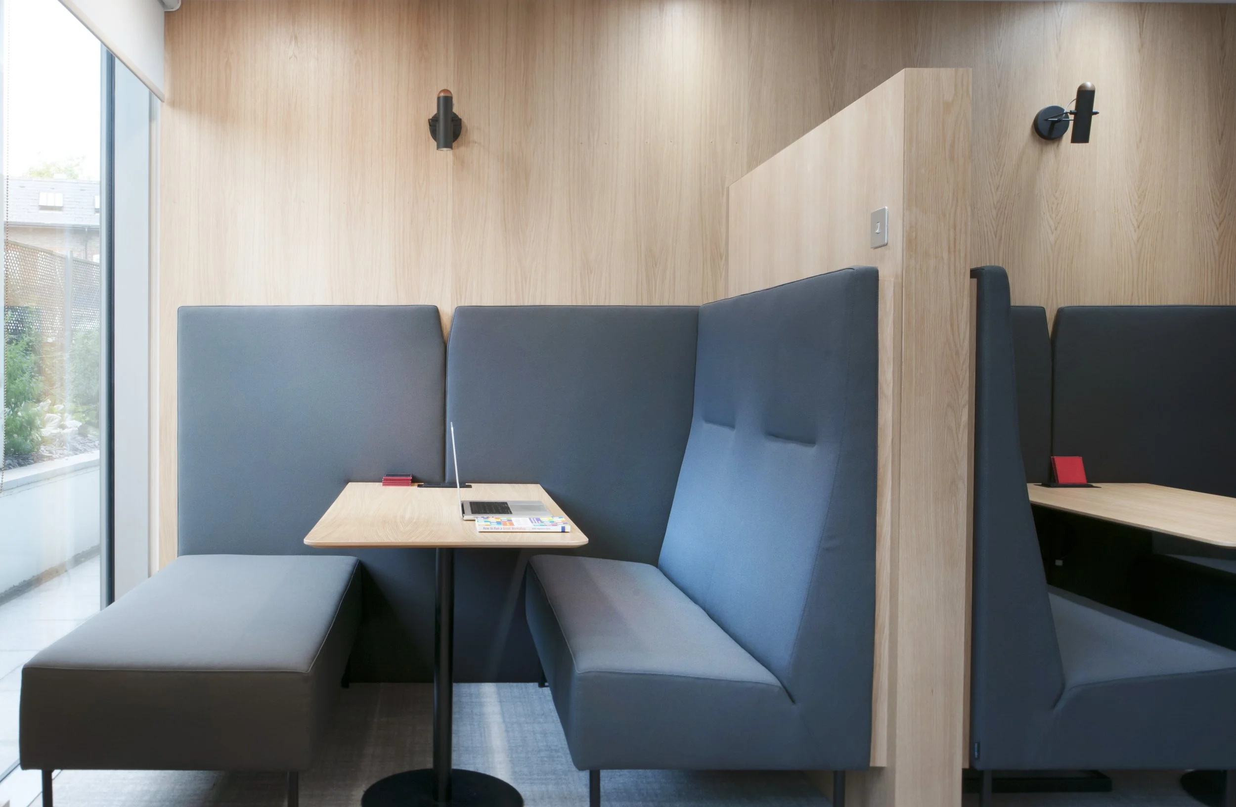 Quiet modern booth seating area with blue upholstered seats and a small wooden table, located next to a floor-to-ceiling window with greenery outside, in a contemporary interior space.