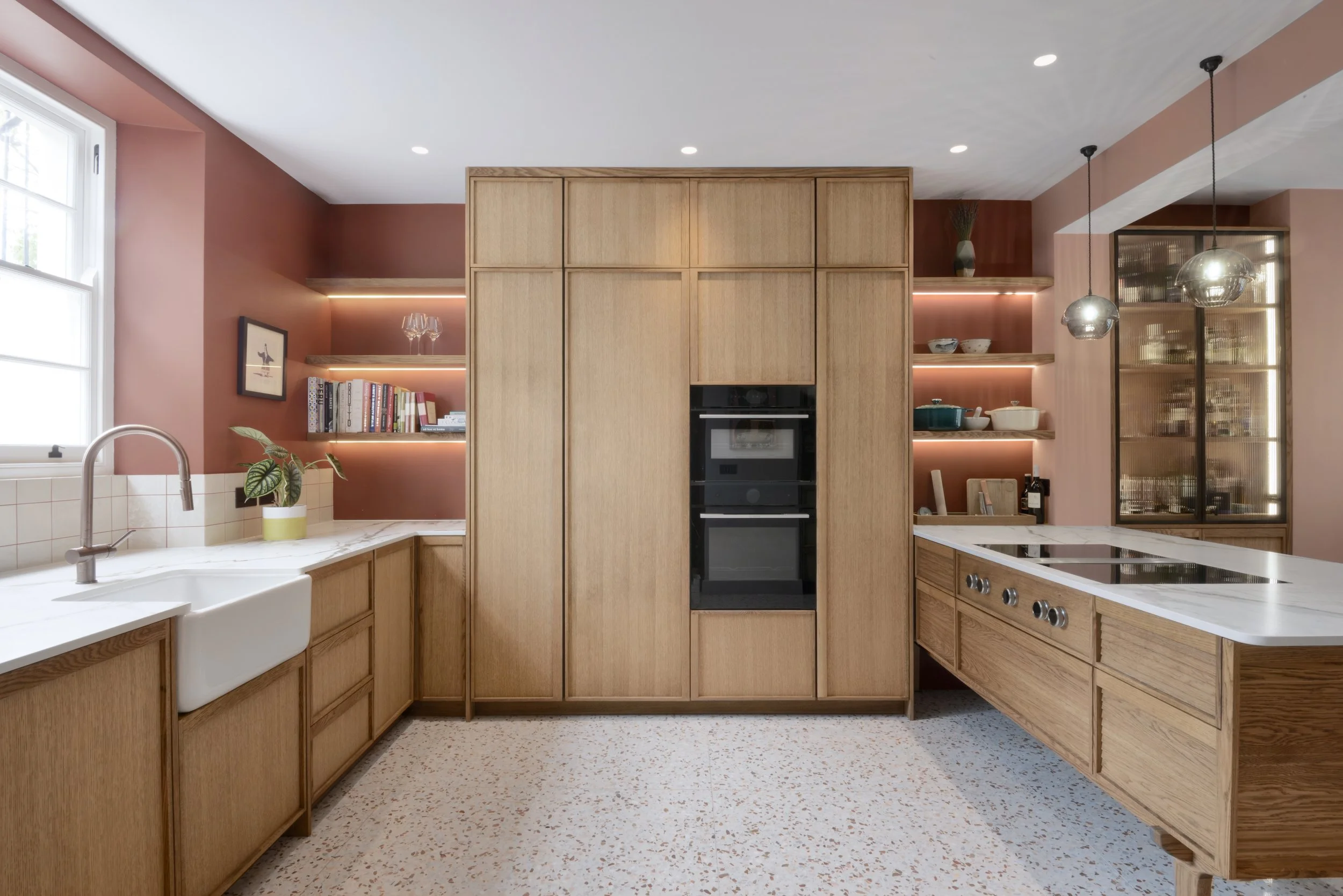 Modern kitchen with wooden cabinets, pink walls, open shelving with book and dishes, a white farmhouse sink, and an induction cooktop on a kitchen island.