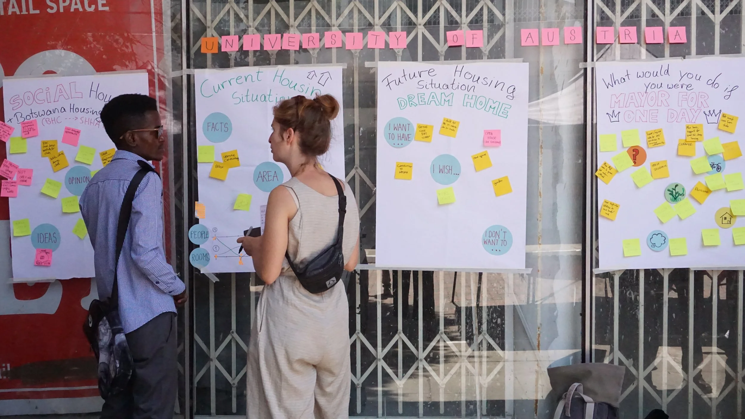 Two people standing and talking in front of large posters about housing options at the University of Austria. The posters contain handwritten notes, post-it notes, and diagrams related to current housing situation, future housing dream home, and opin