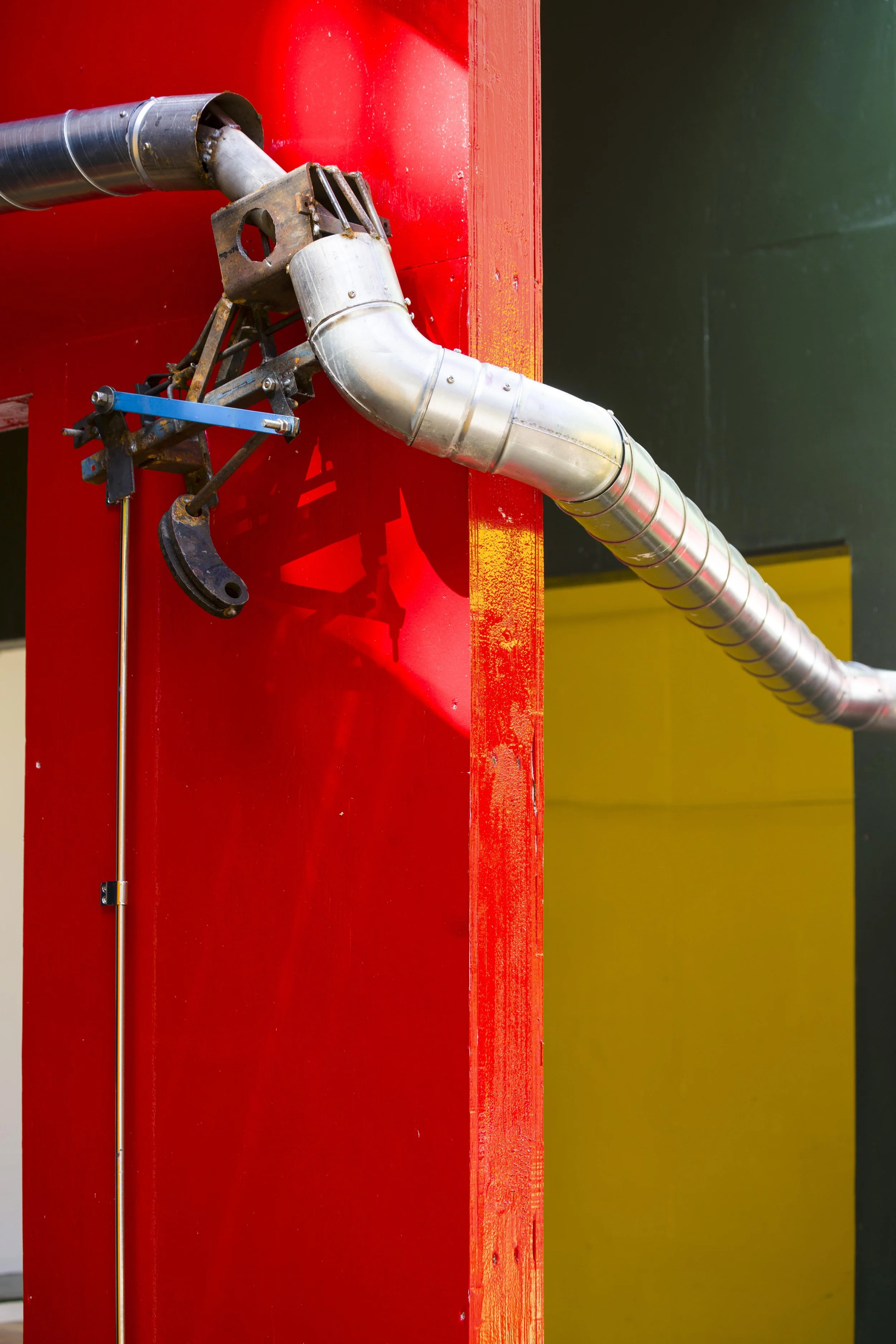 Close-up of industrial metal pipe connected to a red wall, with a yellow section in the background.