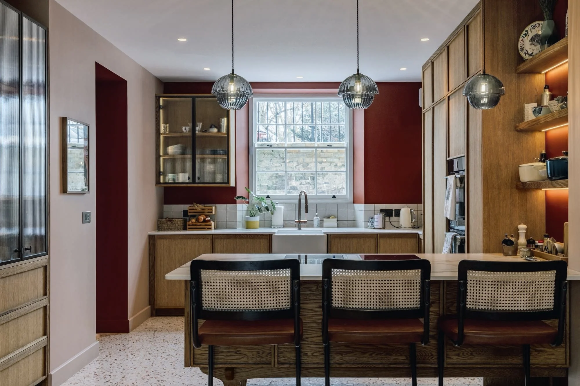A modern kitchen featuring wooden cabinetry, a white farmhouse sink, a window with a stone wall outside, hanging pendant lights, and a breakfast bar with three chairs.