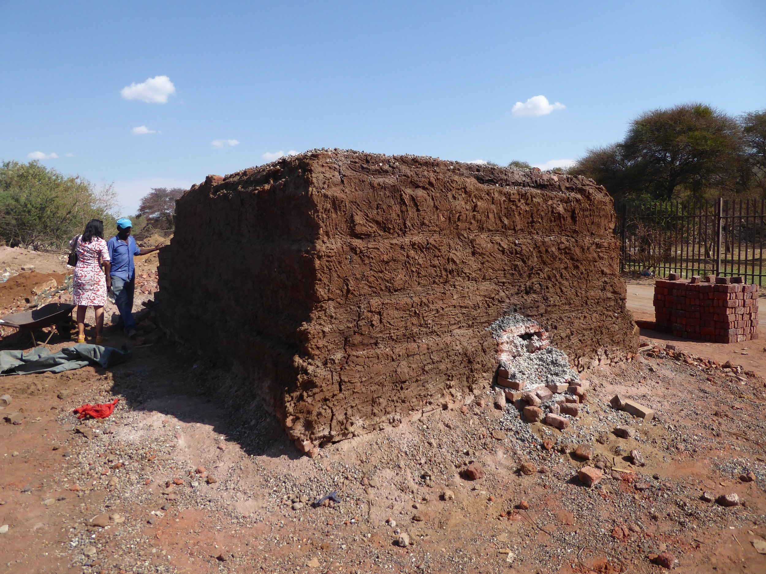 Two people standing next to a large, rectangular, mud-brick structure outdoors with a partly cloudy sky and some trees in the background.