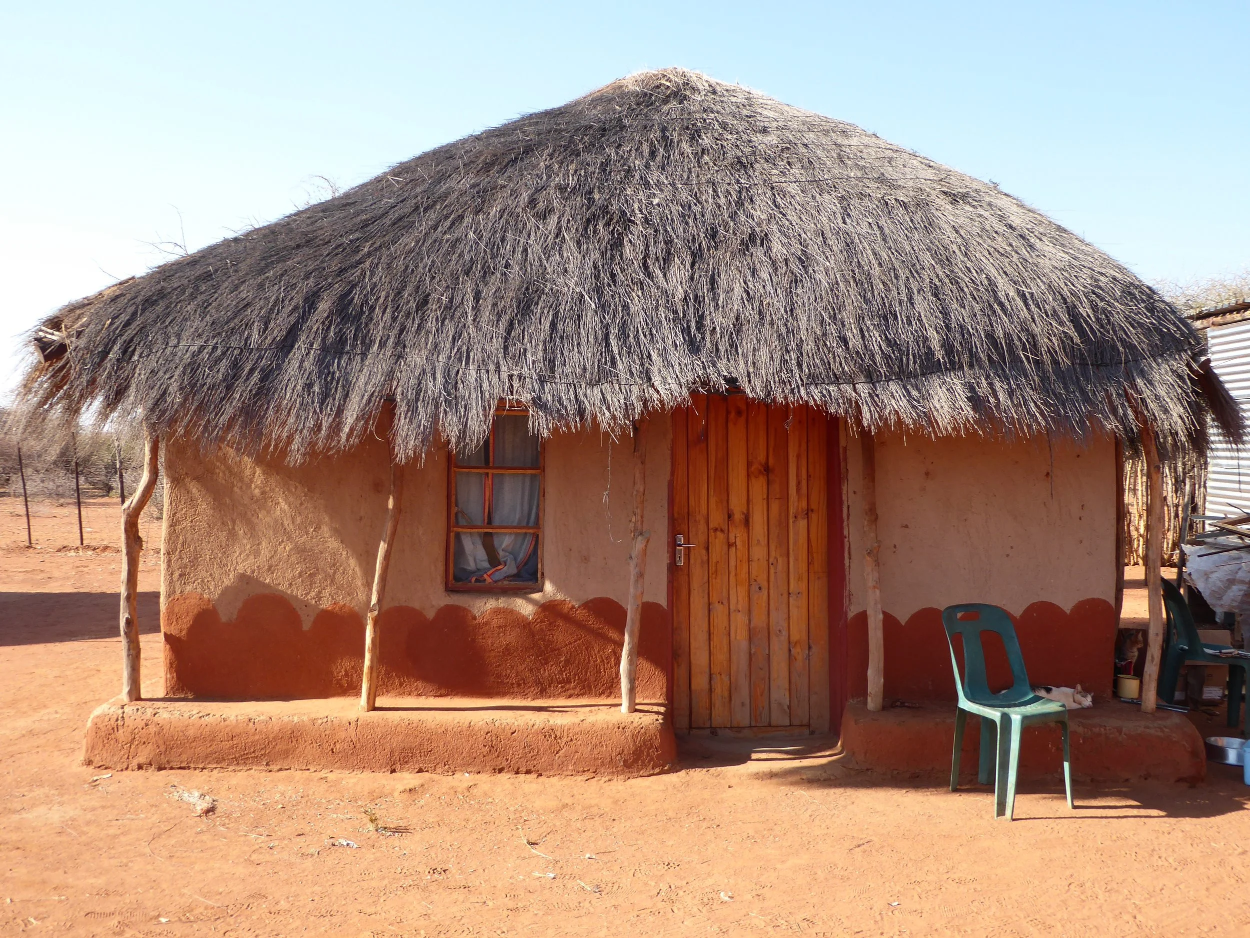 A traditional hut with a thatched roof, mud walls, a wooden door, and a small window, located in a dry, rural area.