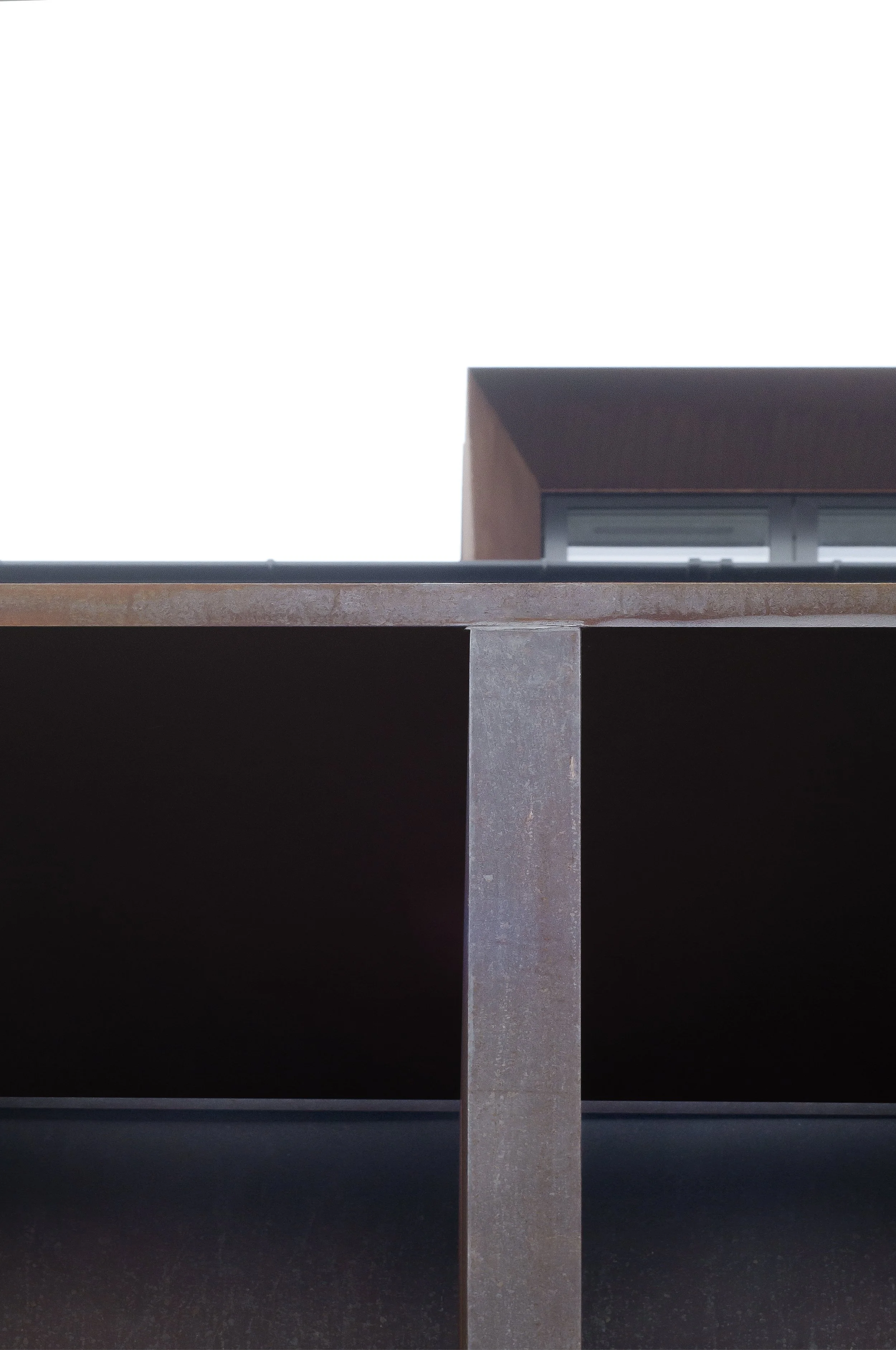 Close-up of a modern building's exterior with metal beams and a large window, against a white sky.