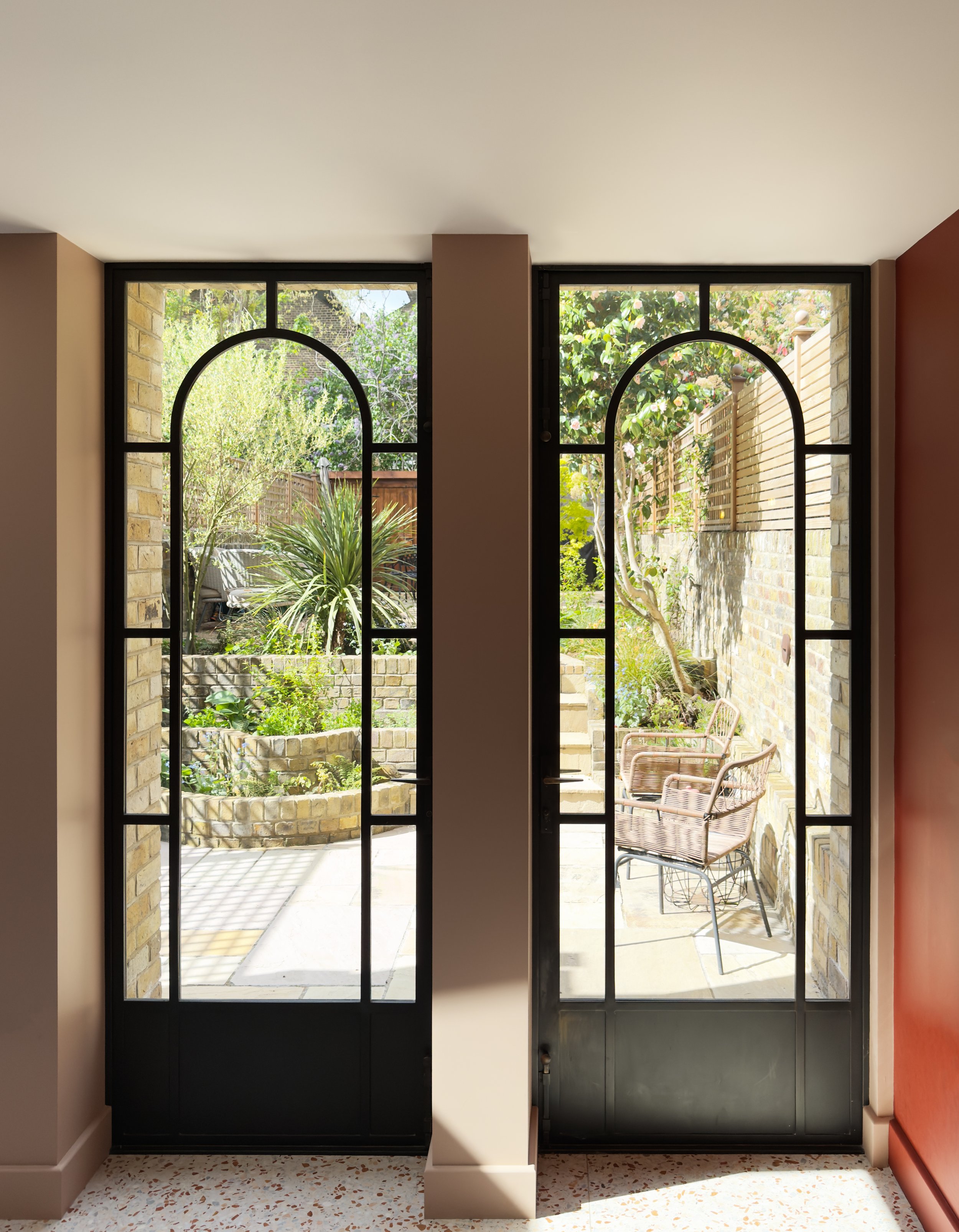 View of a garden through two black framed glass doors with an arched top, showing outdoor plants, patio chairs, brick walls, and a wooden fence.
