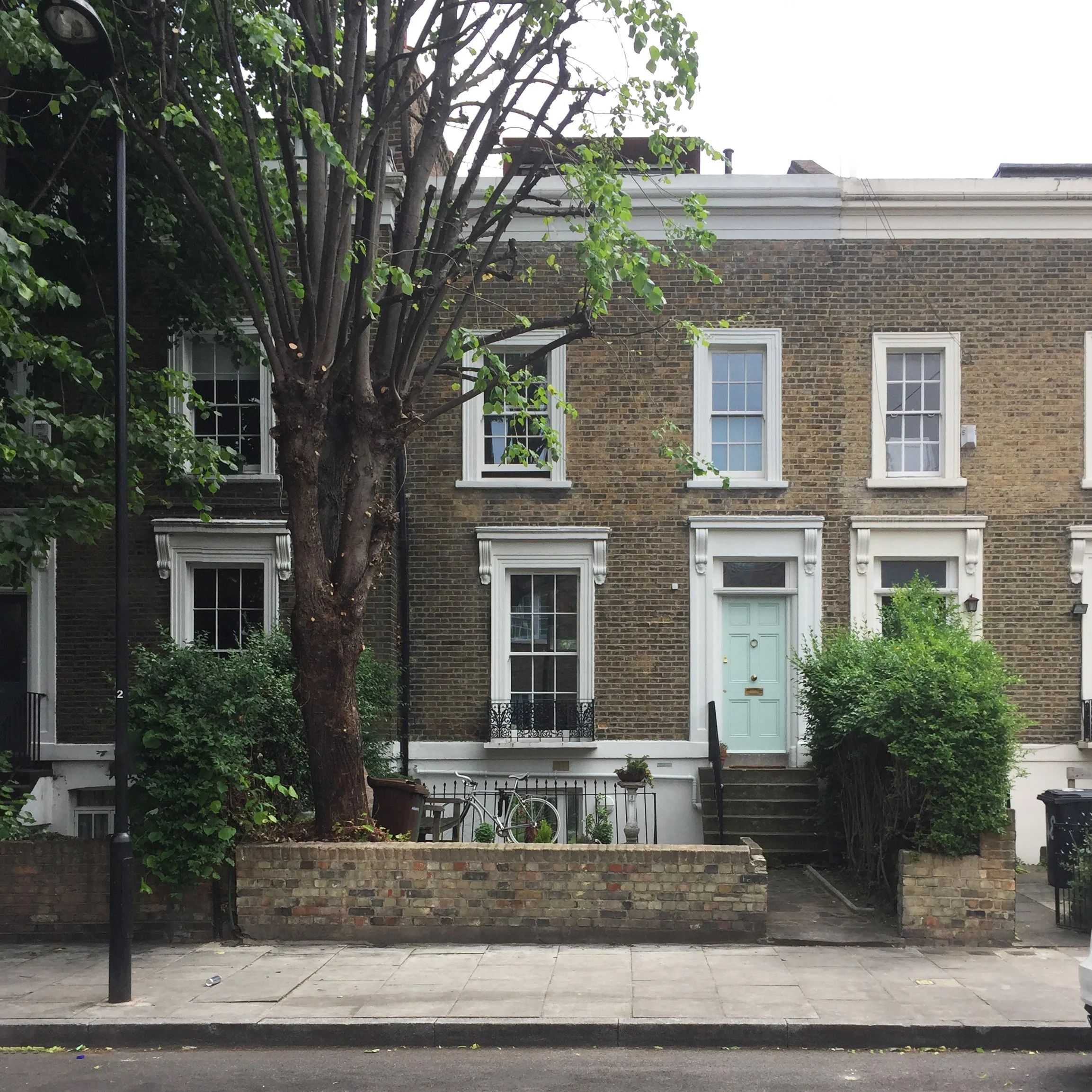 A brick townhouse with white window frames and a mint green front door, a front yard with a tree, bushes, a bicycle, and stairs leading up to the door.