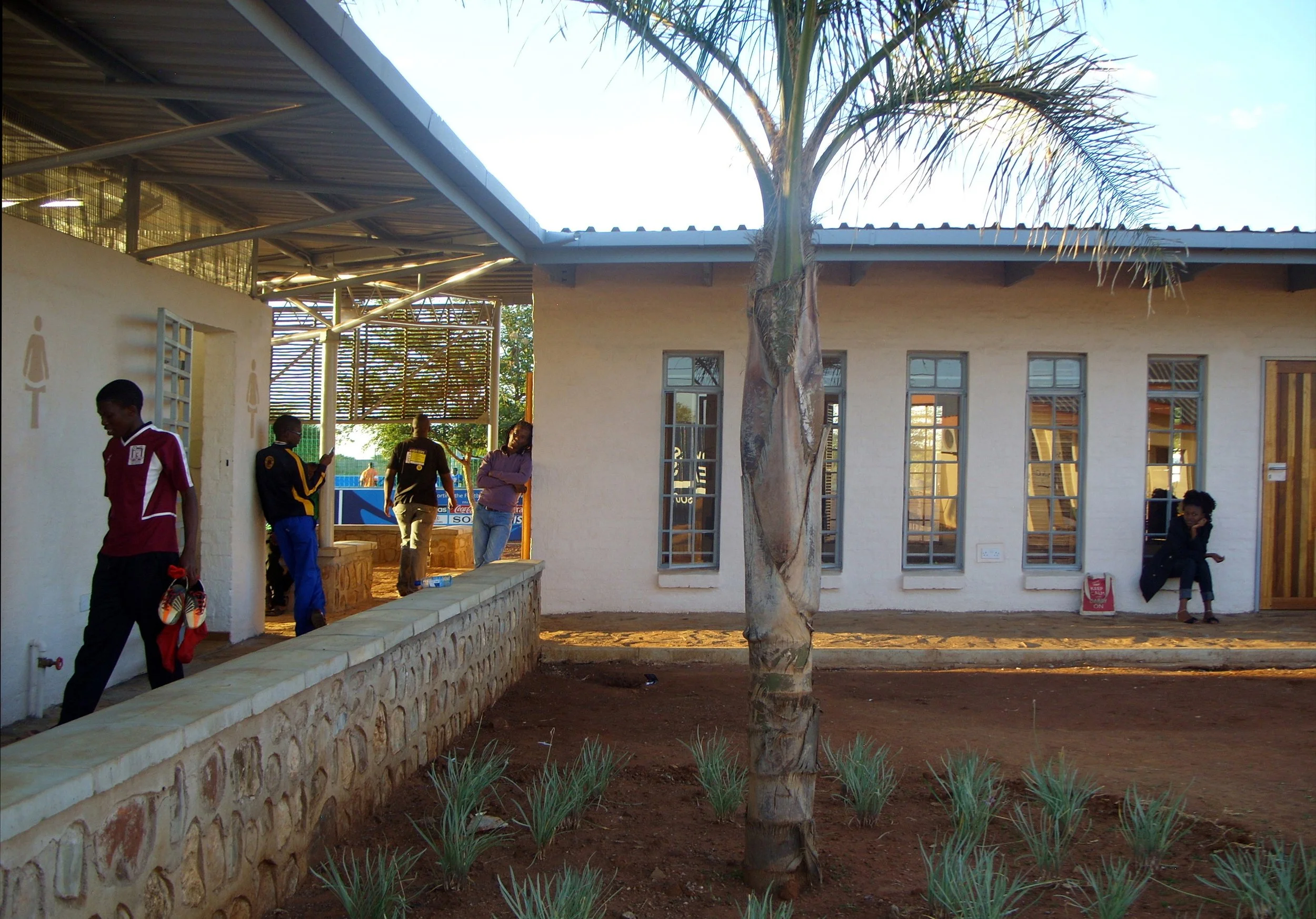 Outdoor scene at a school or community center with a white building, multiple tall windows, and a large palm tree in the foreground. Several people are standing or sitting outside, engaged in conversations or waiting.