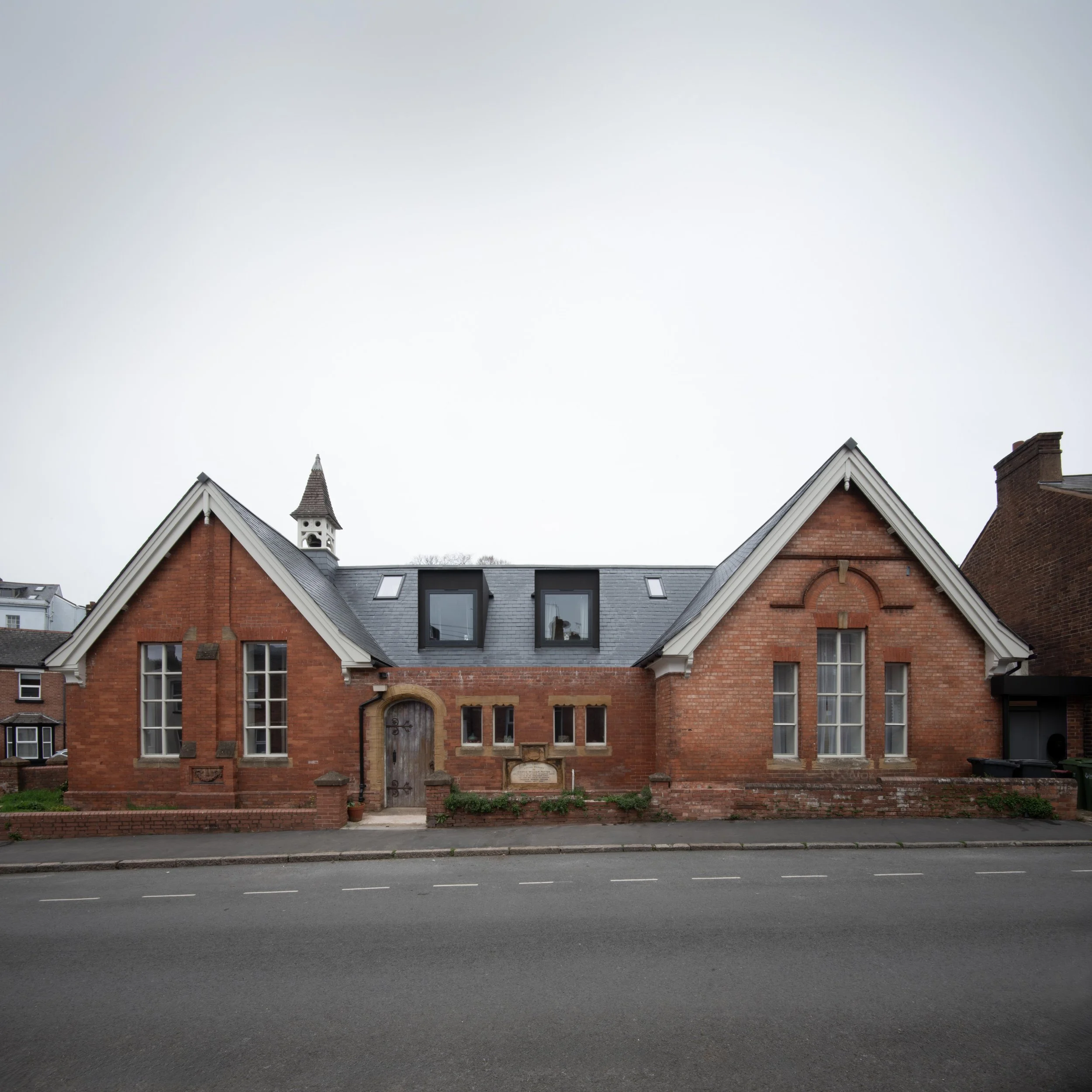 A brick building with artifacts and a plaque near the entrance, located along a street. It has a gabled roof, tall arched windows, and a small steeple in the background.