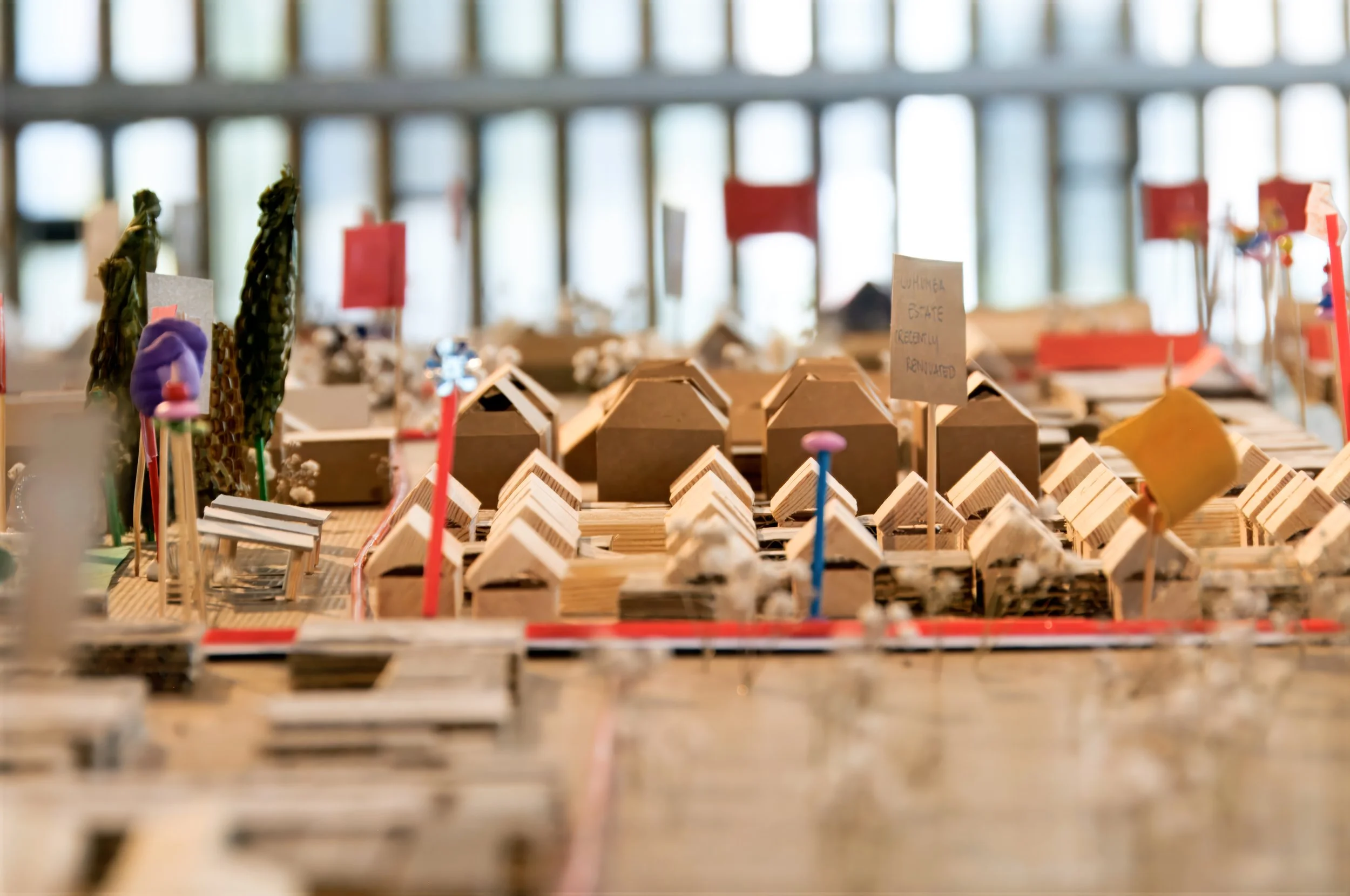 A miniature architectural model with small wooden houses, trees, and colorful flags on a table, representing a planned community or neighborhood development.