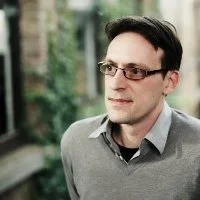 A man with dark hair, glasses, and fair skin wearing a gray sweater stands outdoors in front of a blurred green background.
