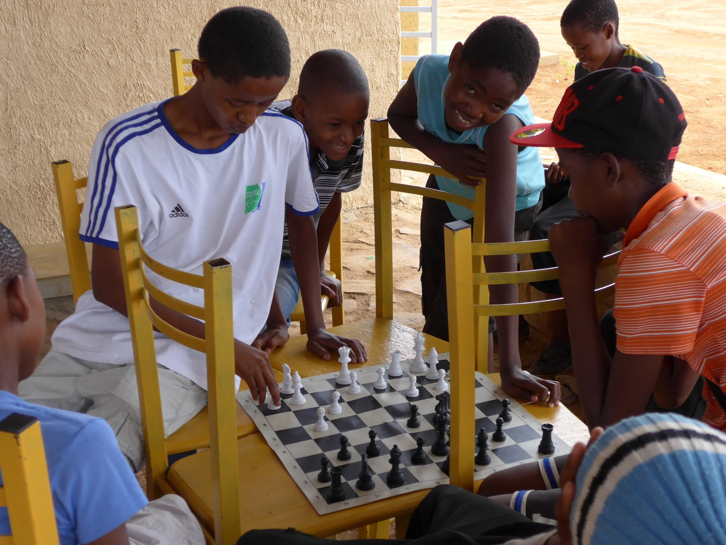 Group of boys playing chess at a wooden table outdoors, with some boys watching and smiling.