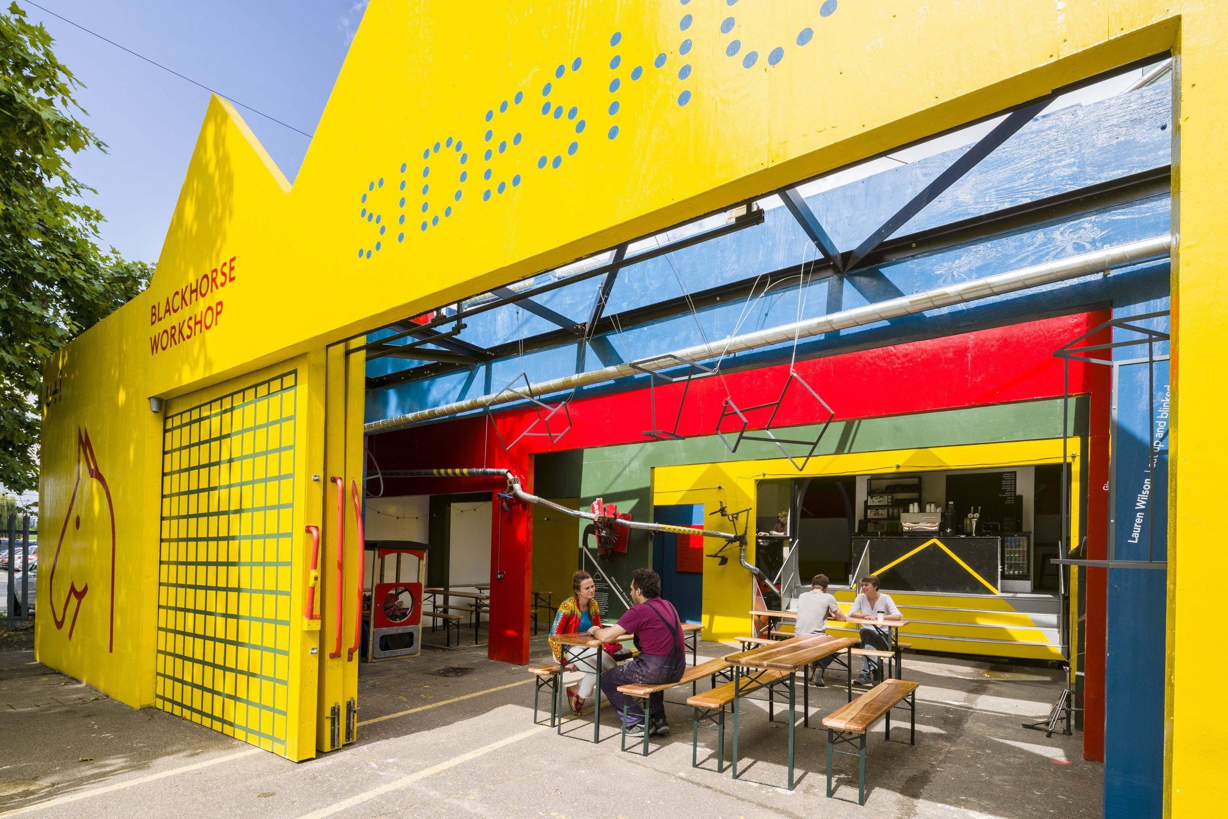 Colorful outdoor installation with a yellow facade, red and green accents, and a semi-open area with picnic tables where people are sitting and talking. The area is shaded with a blue canopy and features artistic and playful design elements.