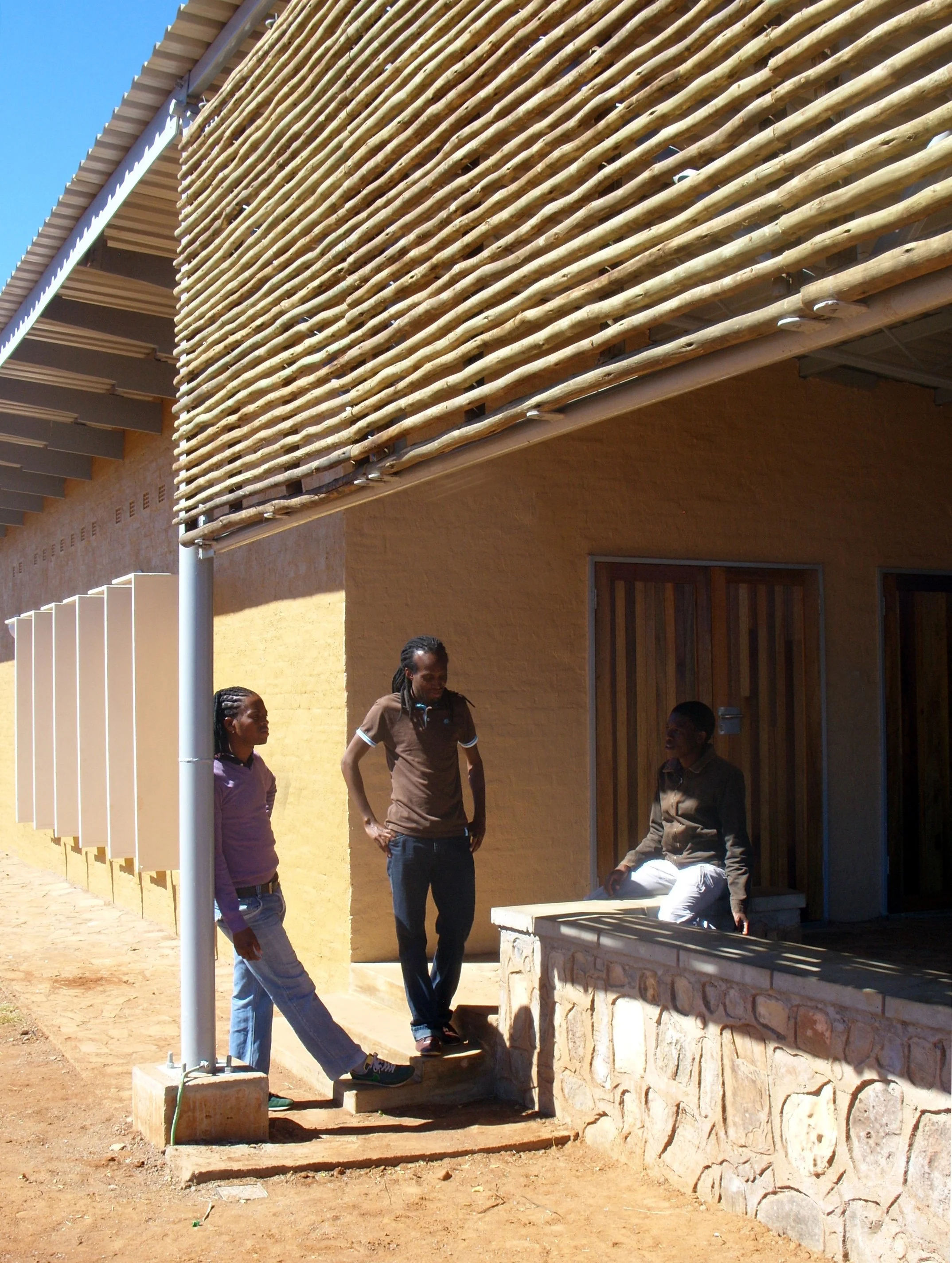 Three women outdoors near a building with a yellow wall, wooden accents, and a decorative wooden slat privacy screen. One woman is sitting on a stone ledge, and two others are standing, engaged in conversation.