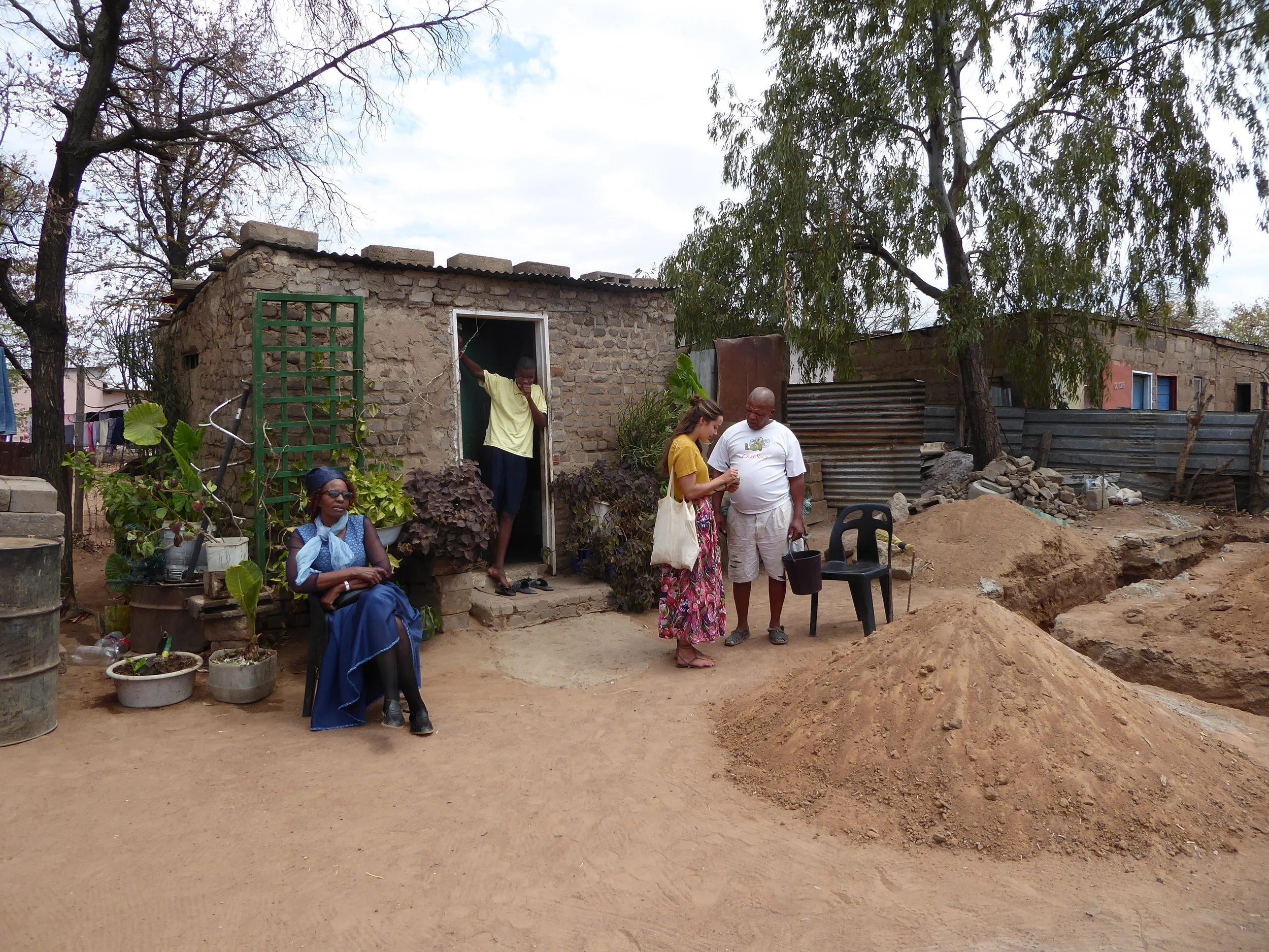 Four people are outside a small, rustic house made of bricks. One person is seated on a plastic chair wearing a blue dress and sunglasses. Two others are standing and looking at a phone, with one holding a black bucket. Another person is in the doorw